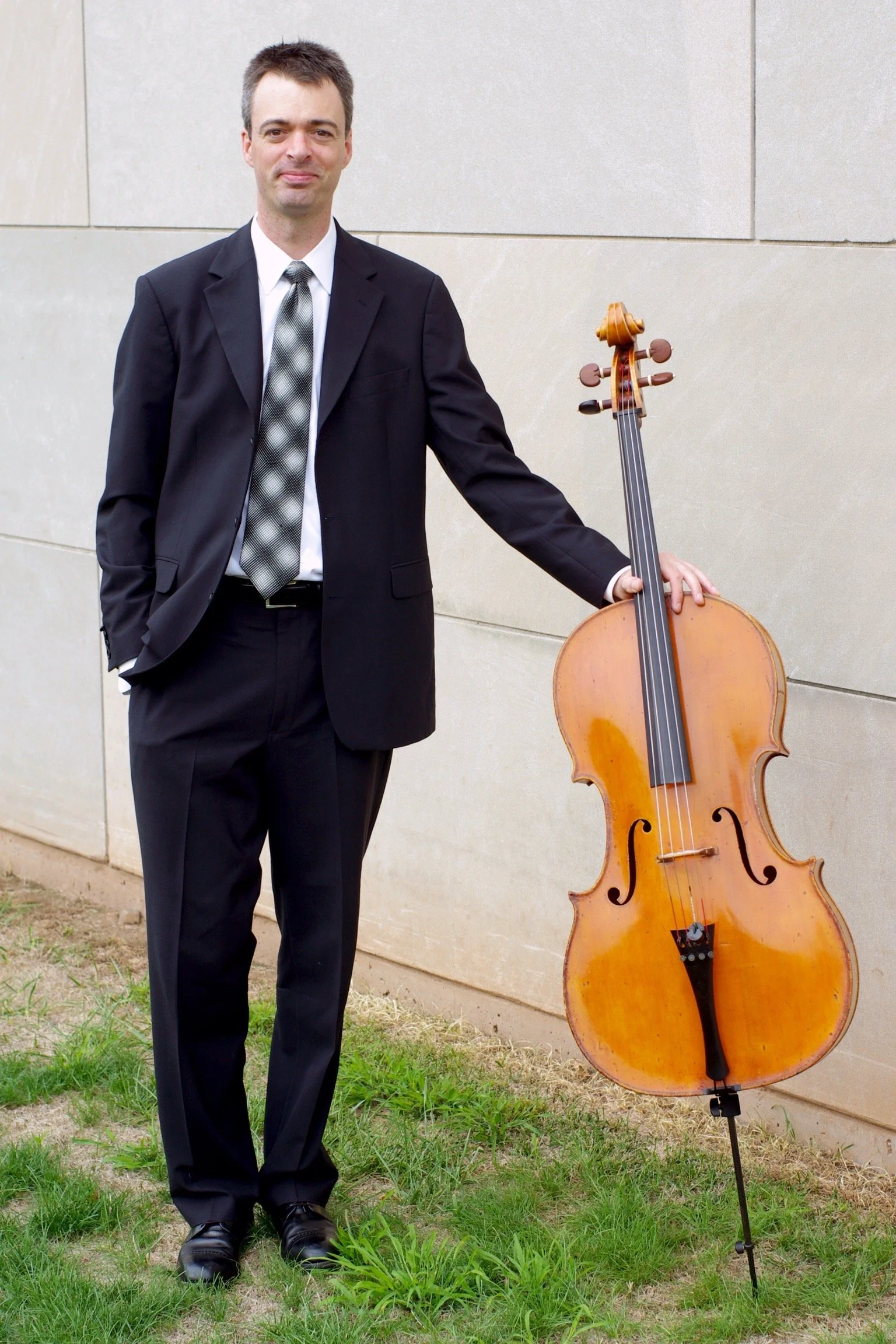 A man in a black suit standing outdoors on grass, holding a cello upright on a stand, against a beige wall.