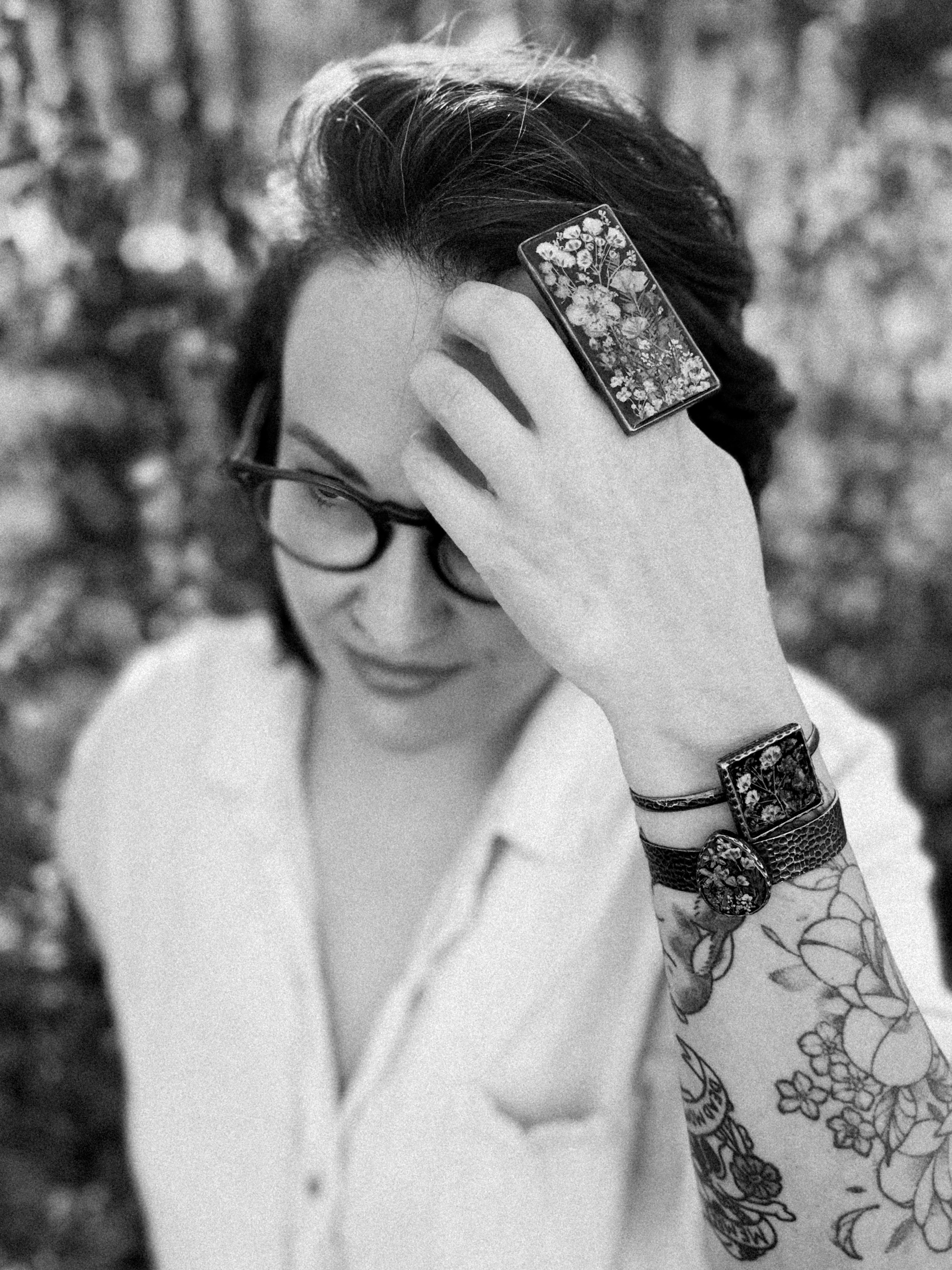 An image of a woman wearing a large ring filled with pressed flowers. She has dark hair and glasses and is running her hand through her hair. She also has on two large bracelets and a white linen button down top. The image is black and white.
