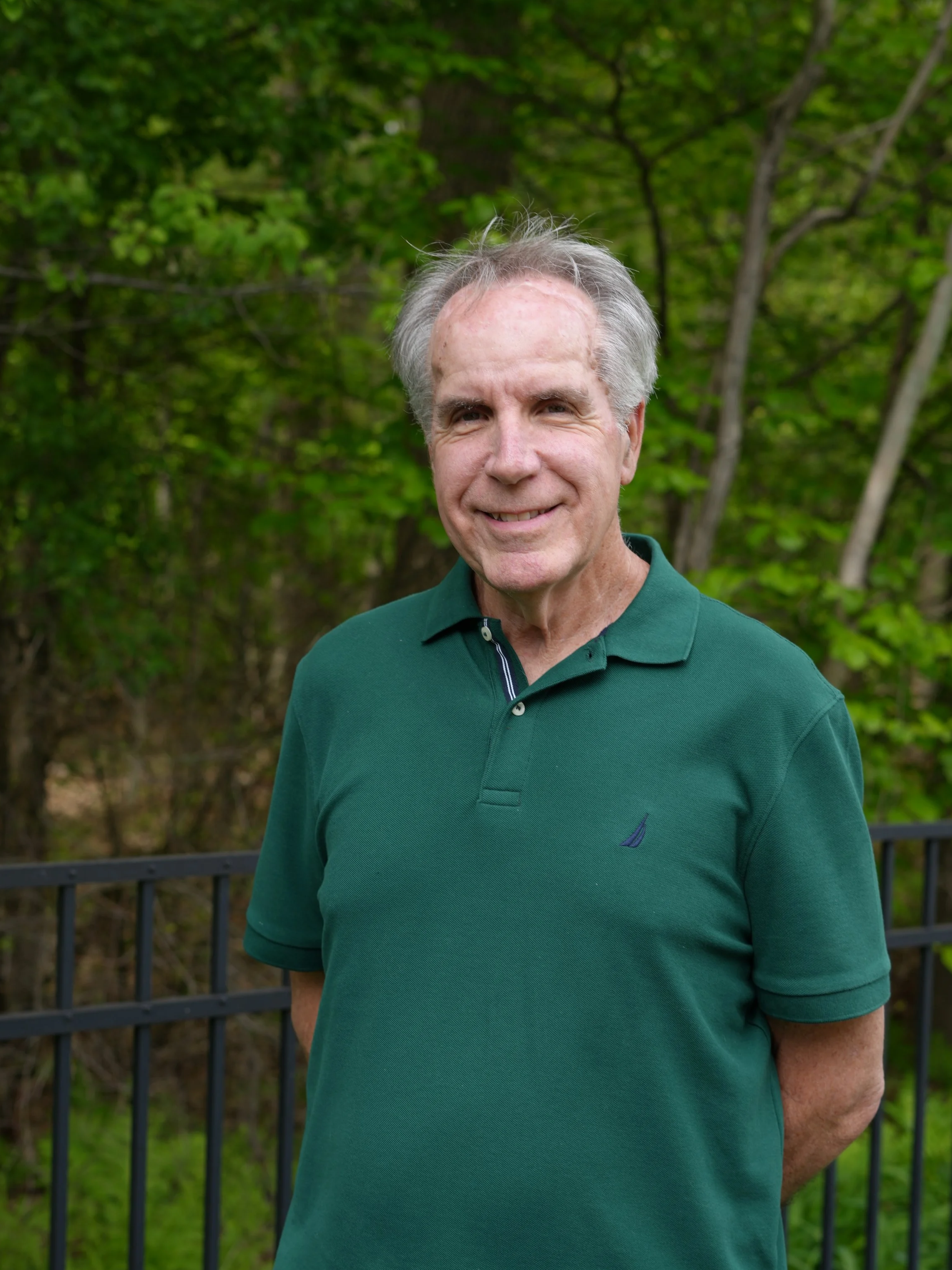 Smiling man wearing a green polo shirt standing outdoors in front of trees and a black metal fence.