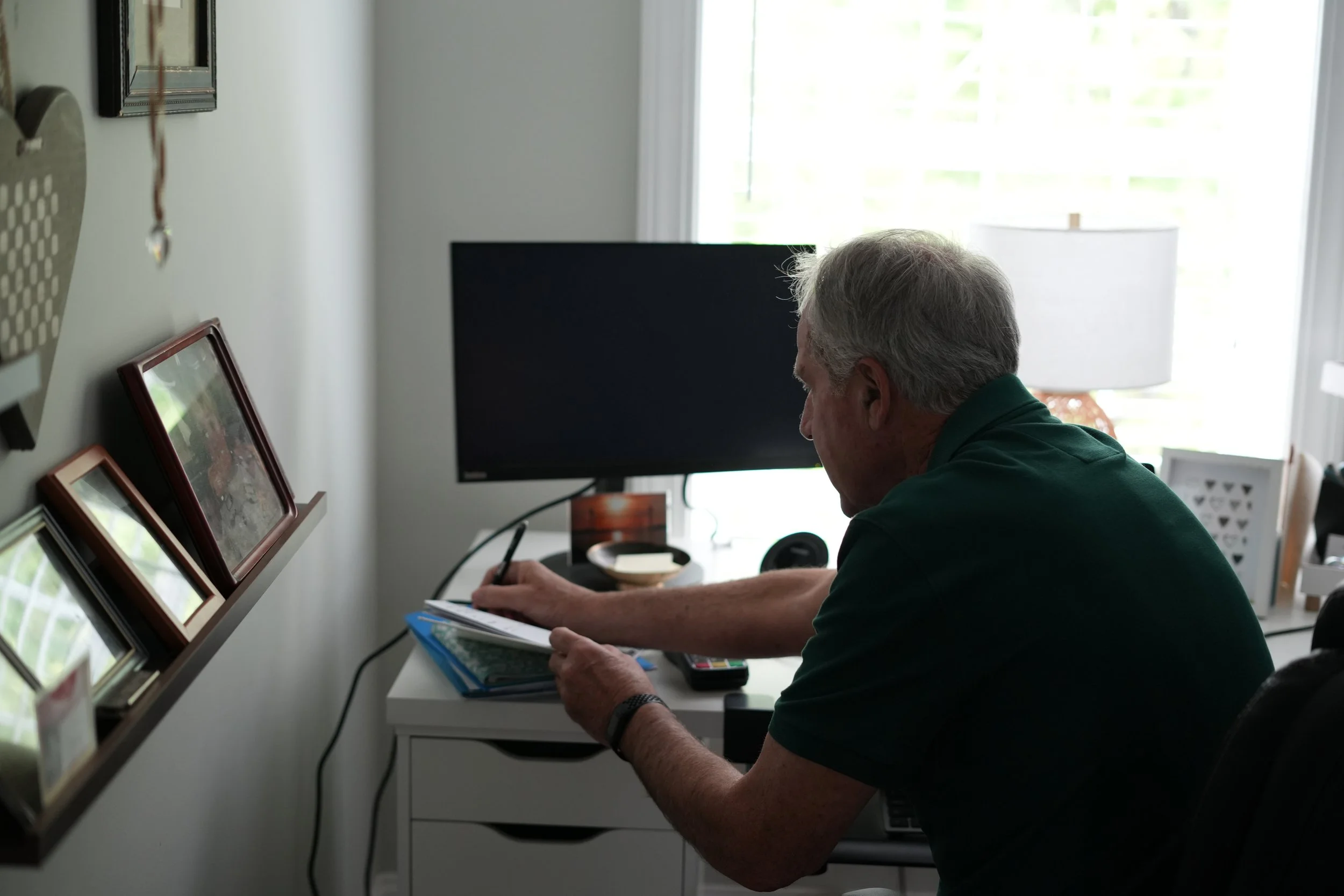 A man sitting at a desk in front of a large window, looking down at a notebook or paper, holding a pen in his right hand, with several framed photographs on the wall behind him and a television or monitor in the background.