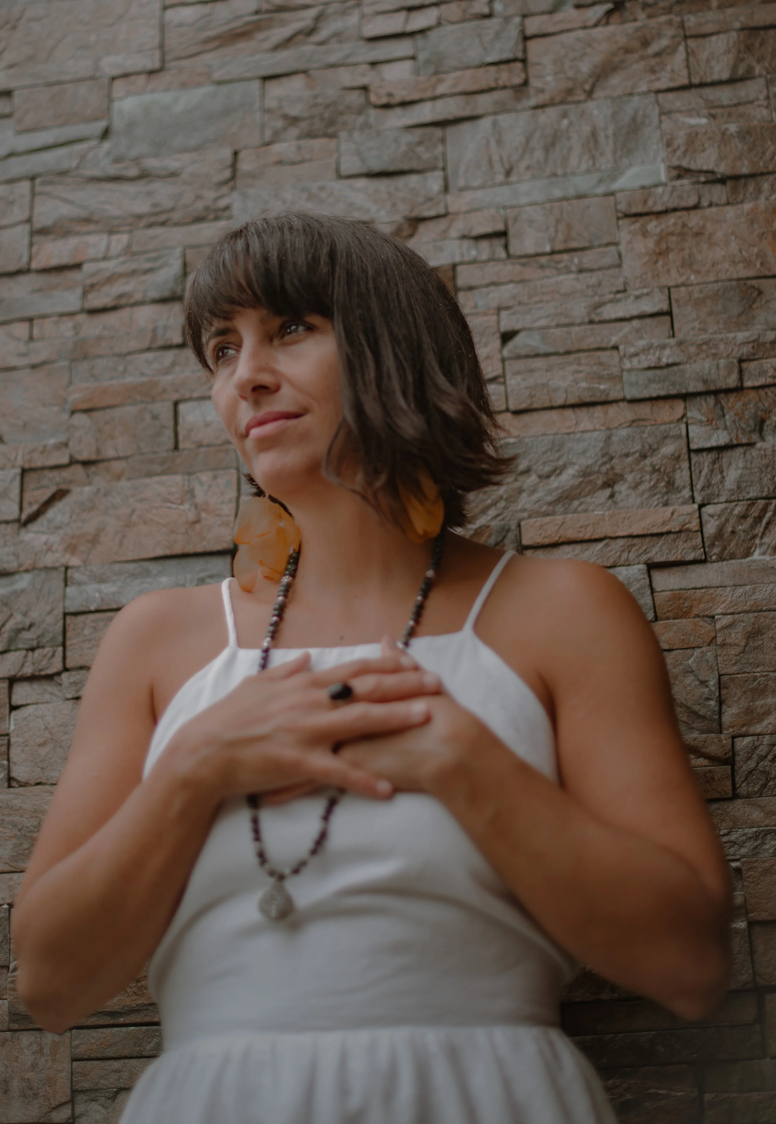 A woman with shoulder-length dark hair, wearing a white spaghetti strap dress, standing in front of a stone brick wall. She has her hands over her chest, wears a beaded necklace and orange earrings, and looks slightly upward with a gentle expression.