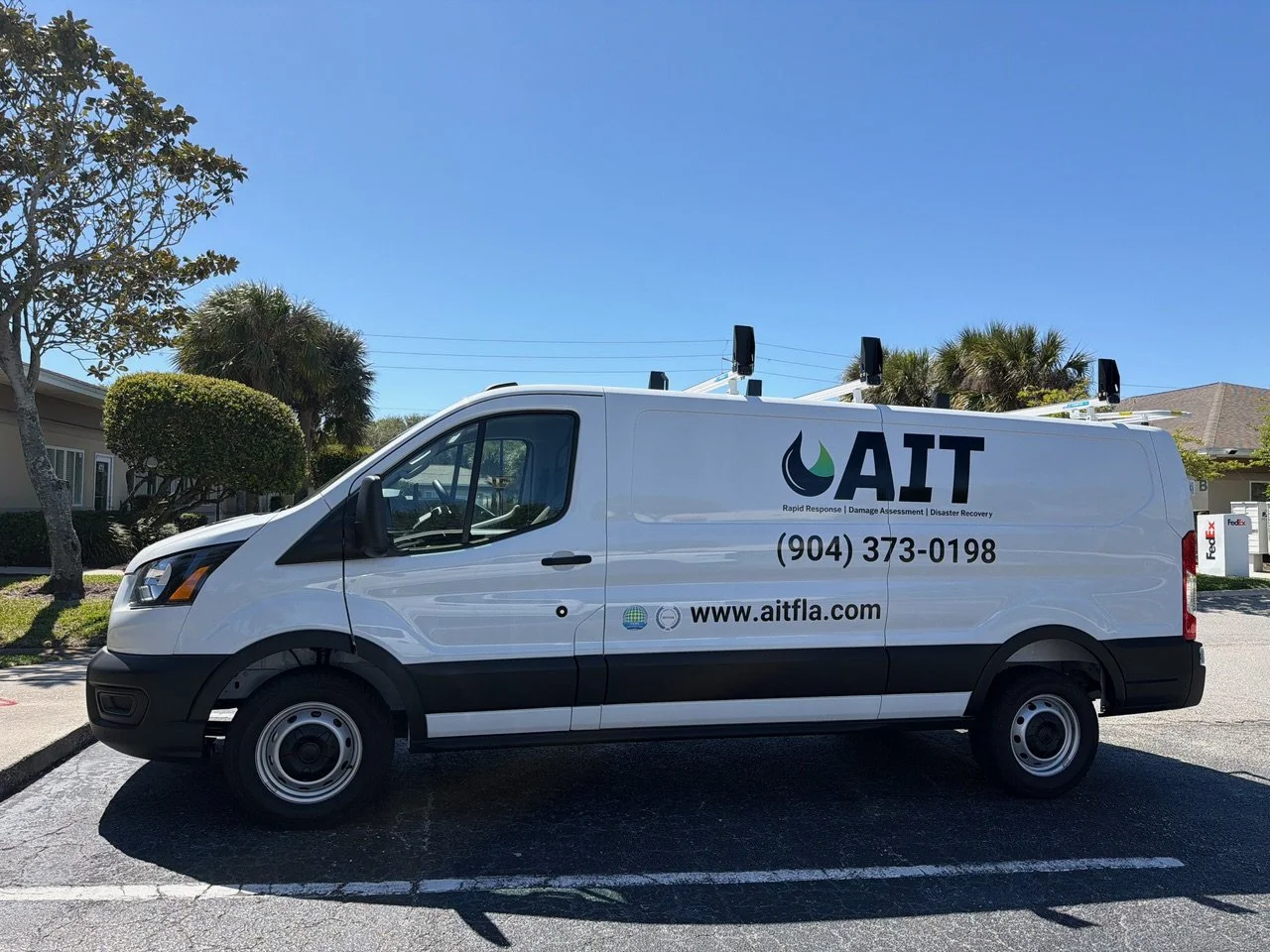 A white AIT service van parked in a lot, with trees and a building in the background. The van has logos and contact information printed on the side, including the website www.aitfla.com.