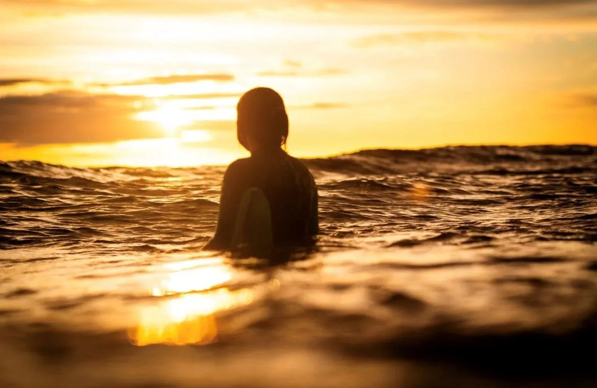 Person sitting in the ocean at sunset with the sun on the horizon and a cloudy sky.