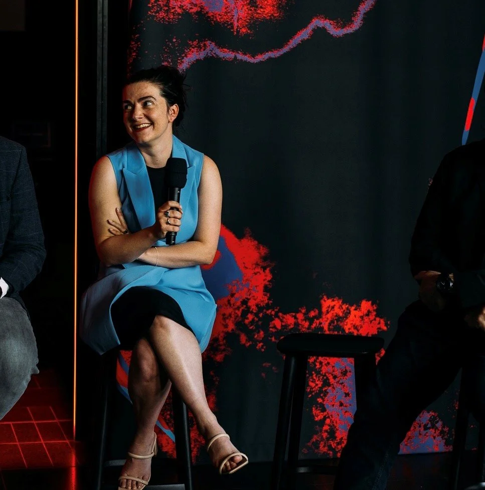 A woman in a blue dress sitting on a stool, holding a microphone, smiling, with a dark background and red and purple abstract patterns.
