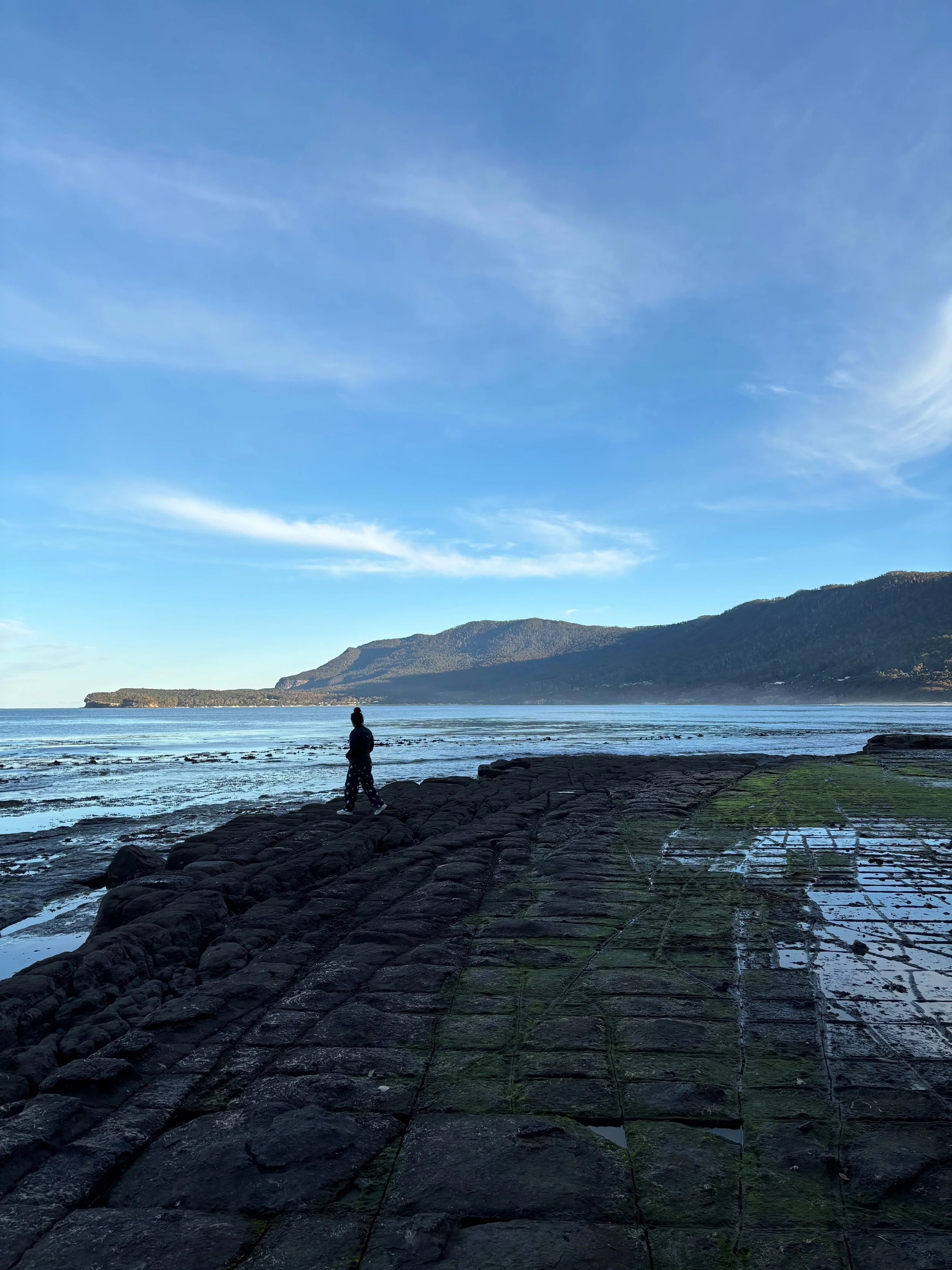 A person walking along a rocky shoreline with water and hills in the background under a blue sky with some wispy clouds.