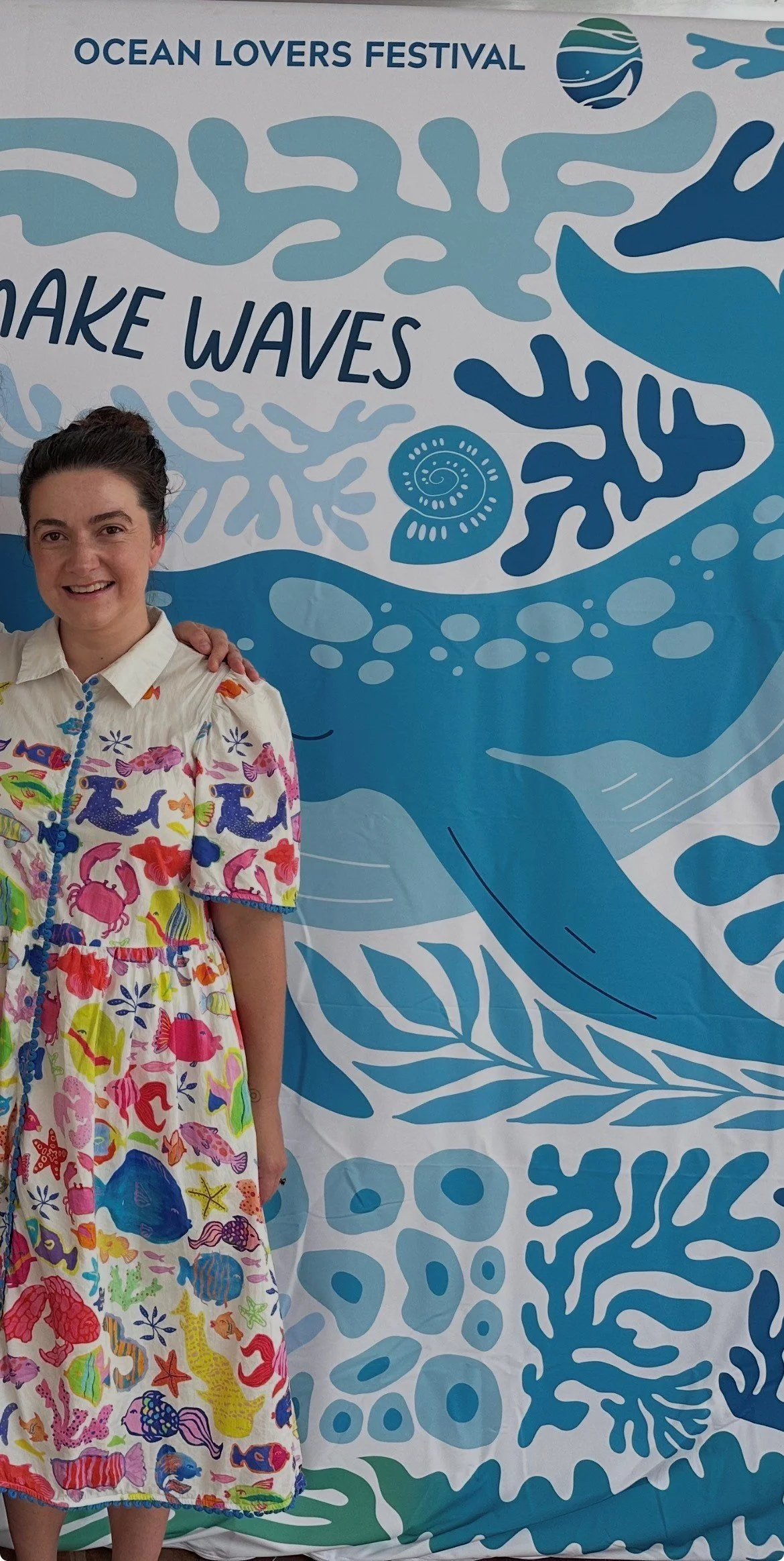 Woman wearing a colorful short-sleeved dress with sea life print, standing in front of a blue and white Ocean Lovers Festival banner with ocean-themed graphics and the words 'Make Waves'.