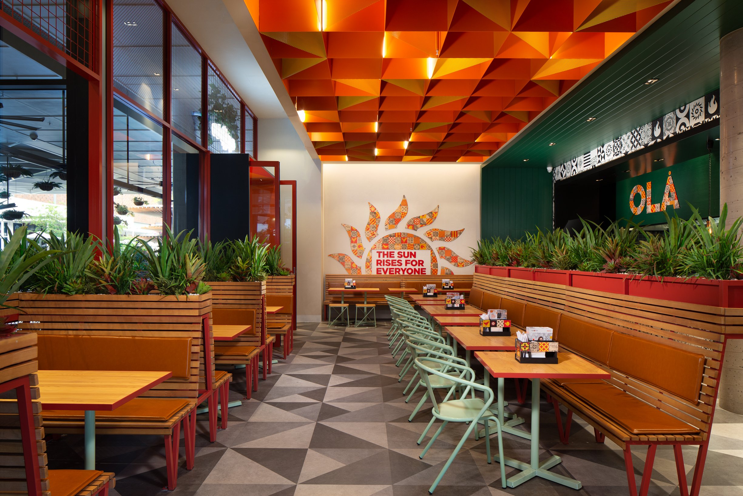 Interior of a modern restaurant with wooden and pastel-colored seating, decorative ceiling panels in warm tones, large windows, and greenery along the perimeter. A wall features a sign that reads "The Sun Rises for Everyone".