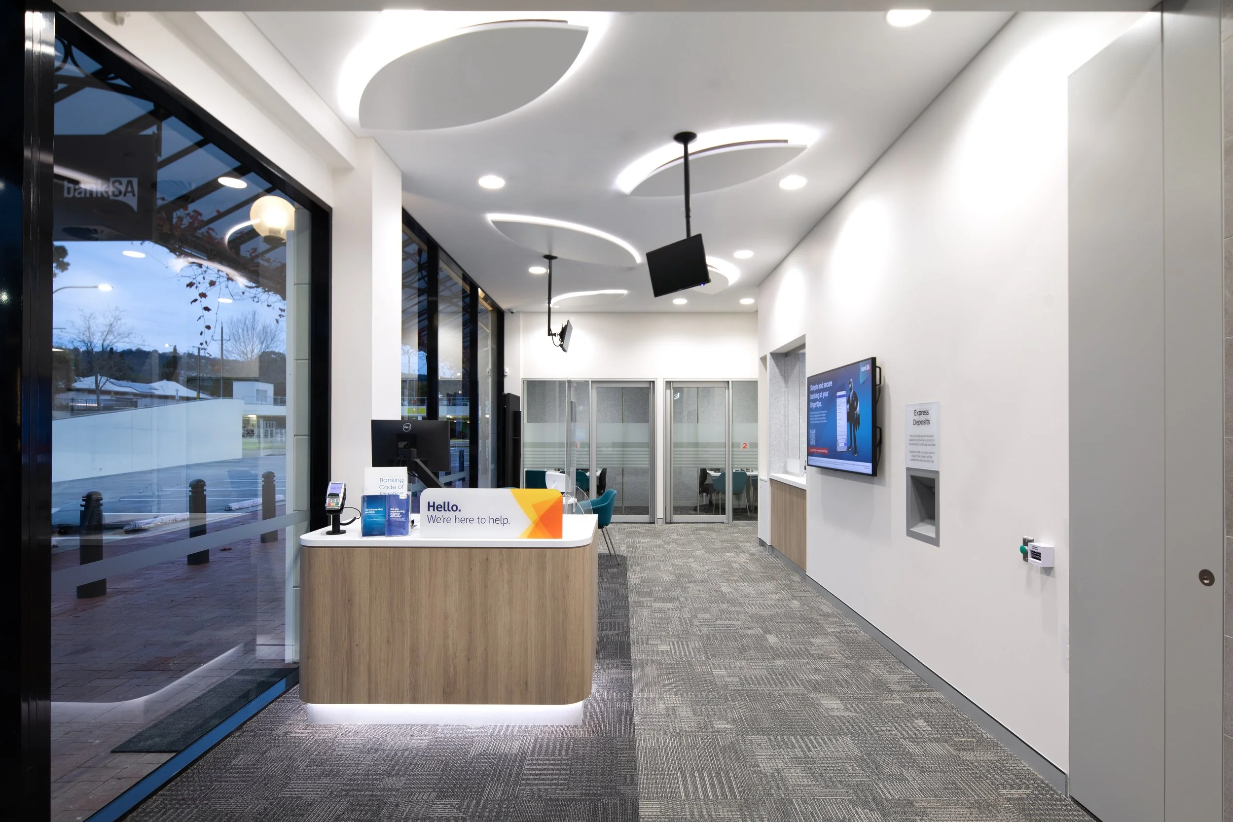 Interior of a modern bank branch with a front desk that has a sign reading 'Hello. We're here to help.' Large glass windows on the left showing an outdoor scene with trees and a building. Ceiling with contemporary curved light fixtures. Wall-mounted 