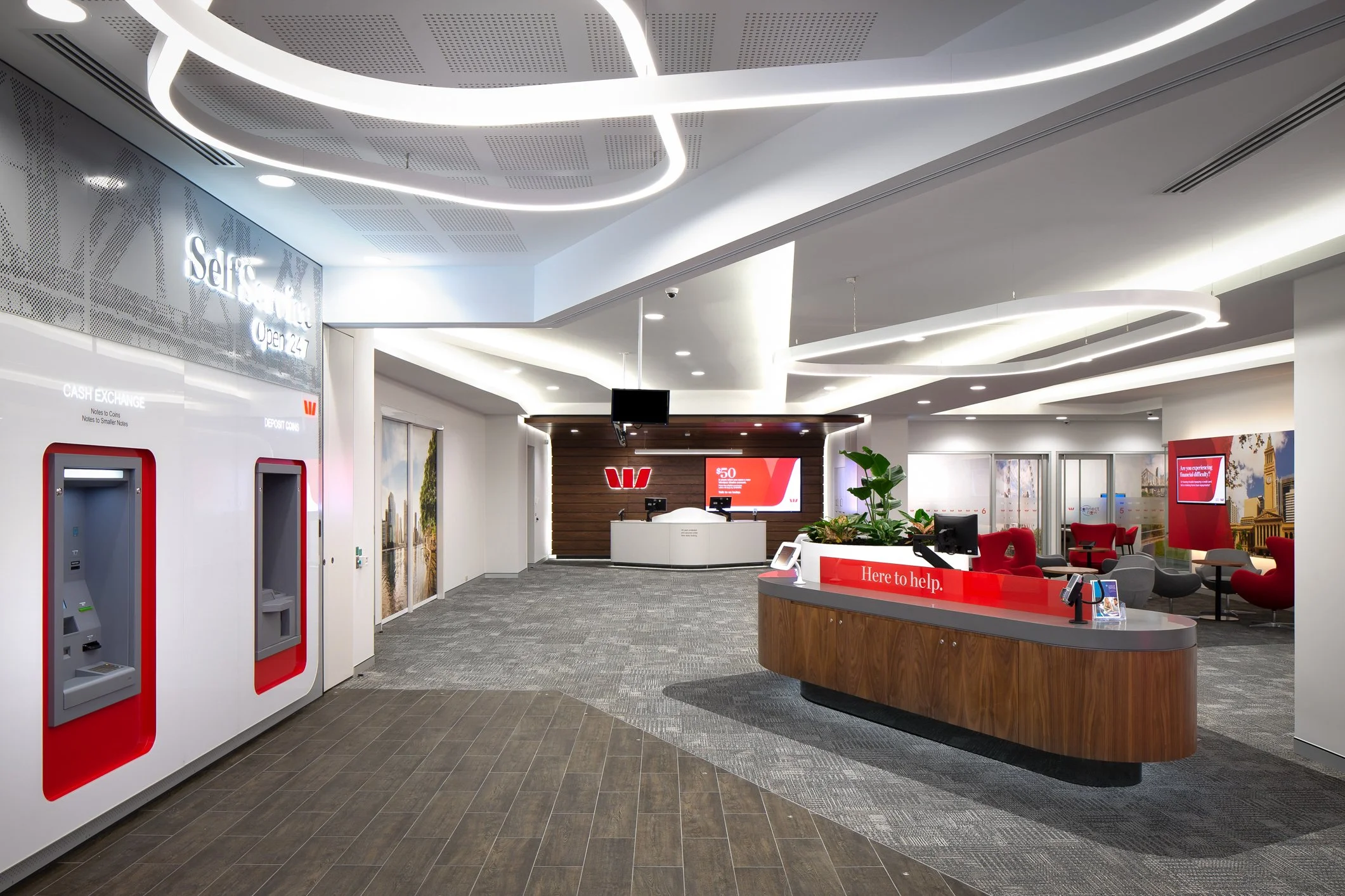 Interior of a modern bank or financial institution with digital teller machines, a reception desk with a red sign that says 'Here to help,' and seating areas with red and gray chairs. The ceiling has illuminated circular and linear light fixtures.