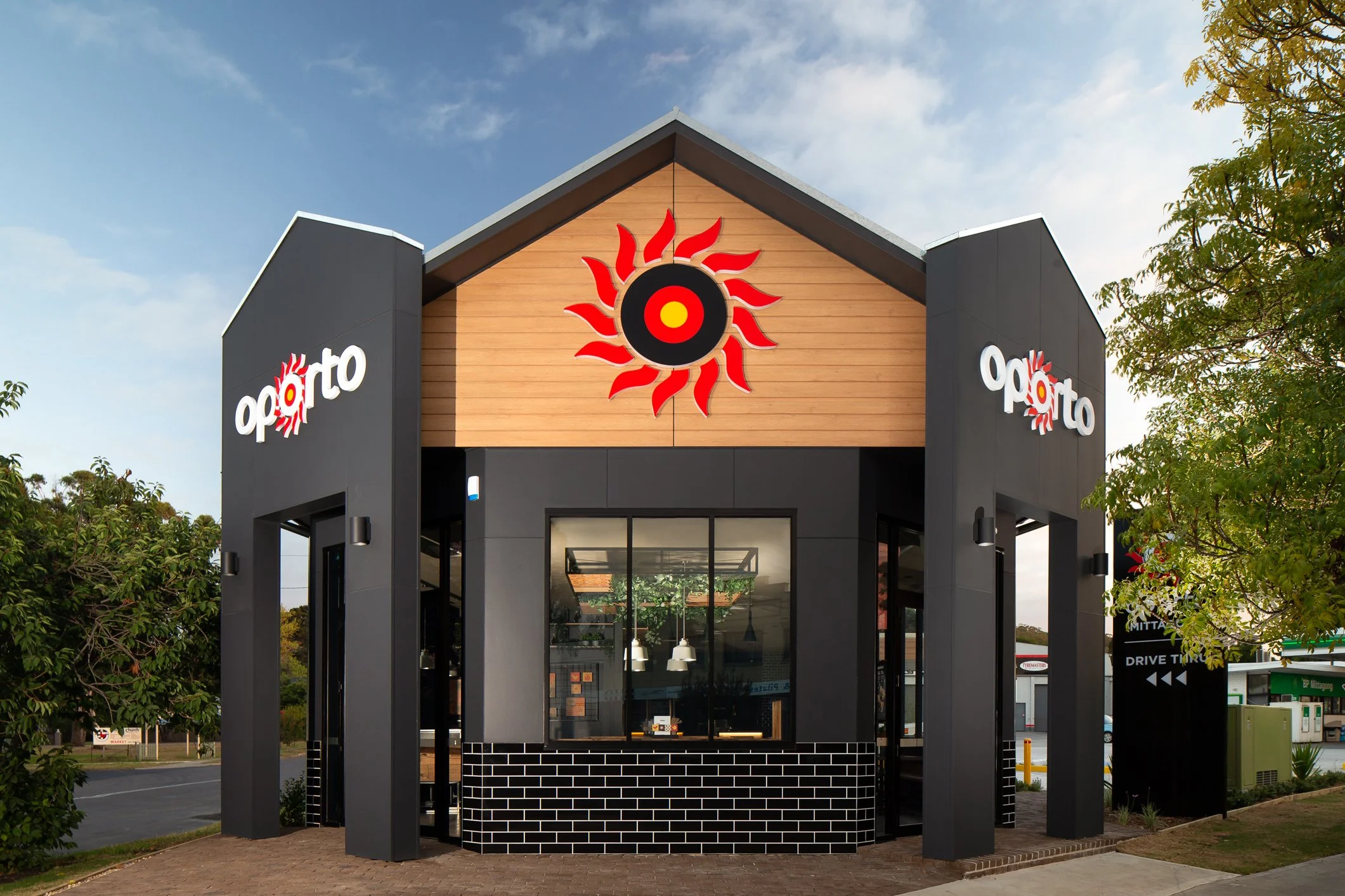 Fast food restaurant building with a modern black and wood facade, featuring a prominent red and black sunburst logo and the name "oporto" on the walls.
