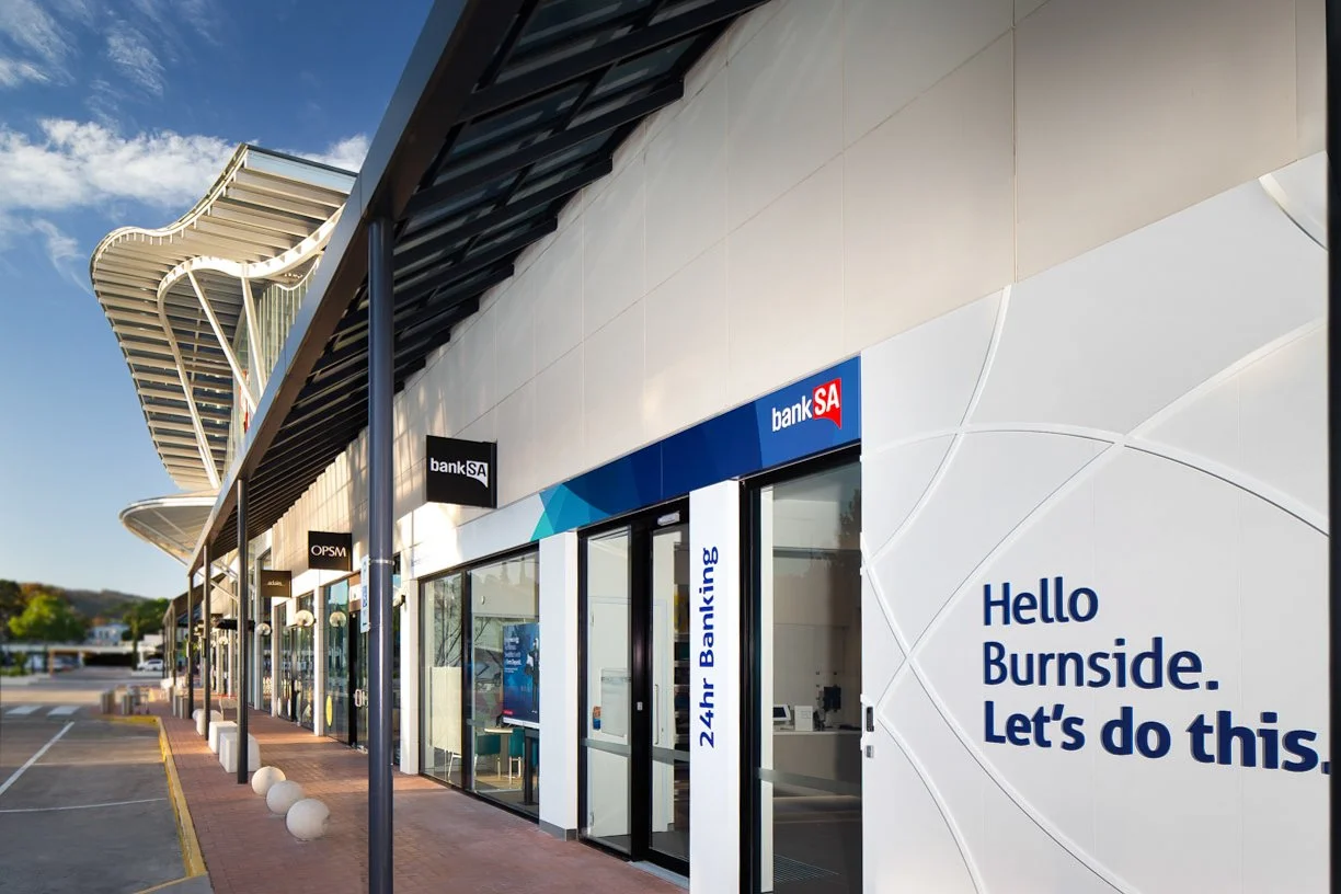 Exterior of a modern banking branch with signage for bankSA and OPSM, and a sign that says 'Hello Burnside. Let's do this.' during daytime.