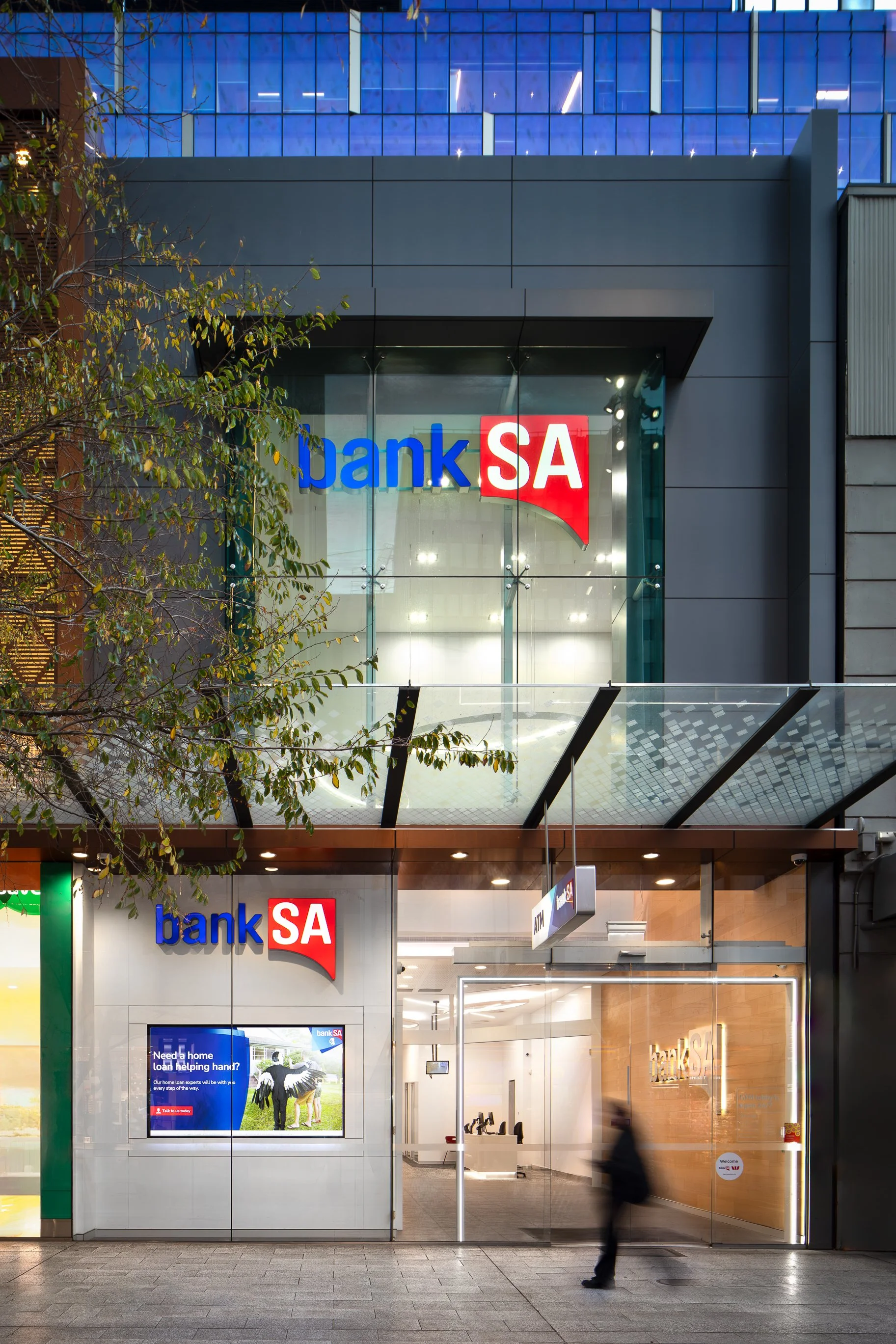 Front view of a modern bank building with a glass facade displaying the 'banka SA' logo in red and blue. Inside, a digital screen shows a message about home loans, and a small staircase and office area are visible.