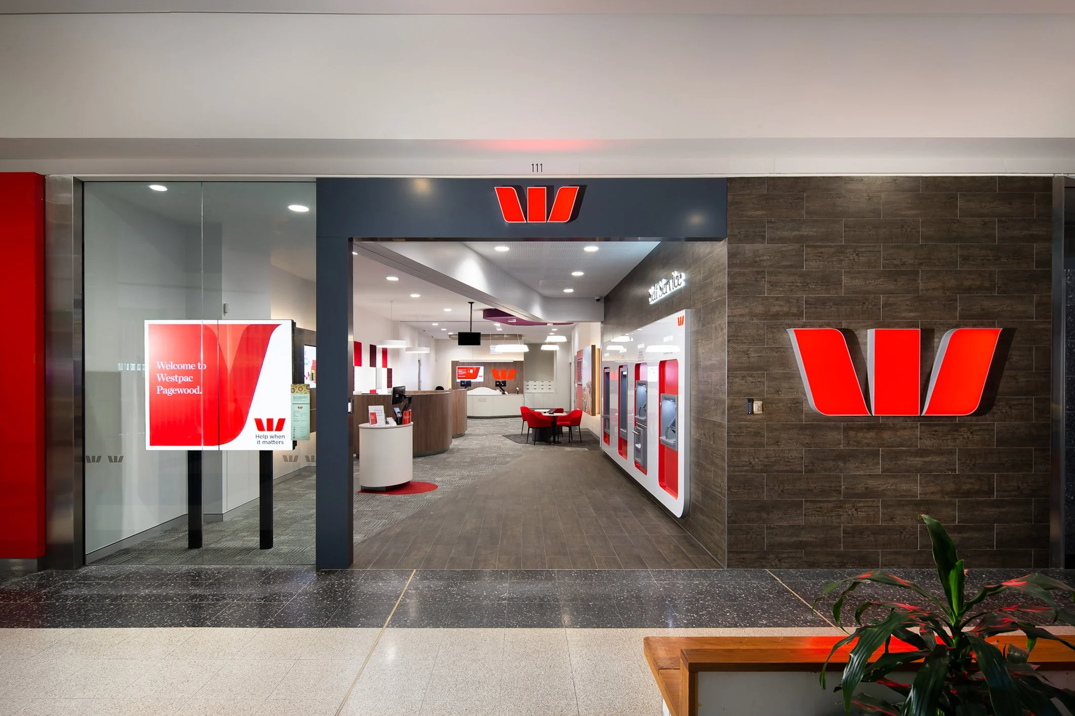 The entrance to a Westpac bank branch with a red Westpac logo above the doorway, glass doors, and digital screens inside displaying messages and advertisements.