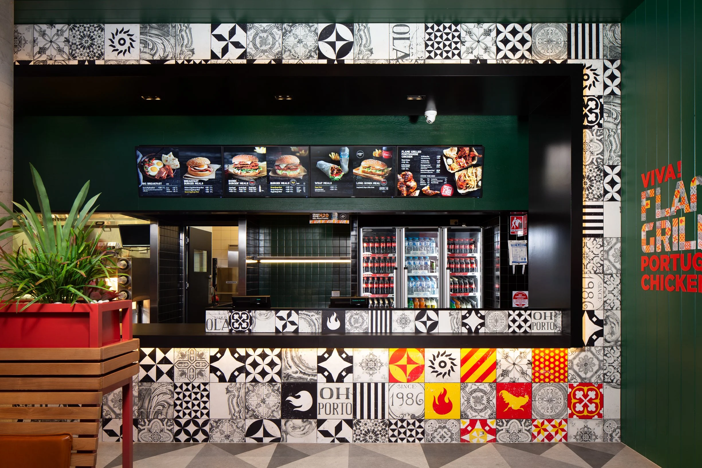 Inside a fast-food restaurant with a decorative tiled wall, digital menu boards displaying burgers and meals, a refrigerator stocked with drinks, and a decorative green wall with a red plant box in the foreground.