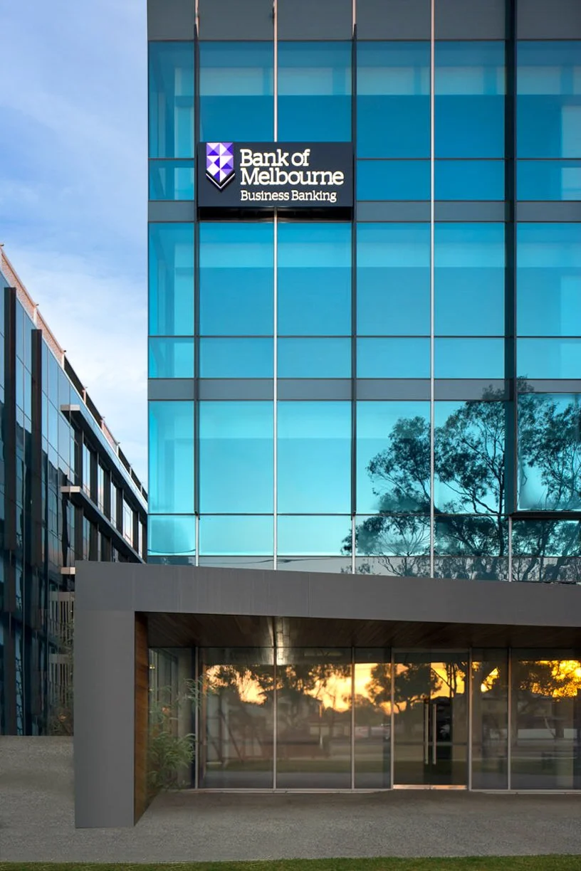 Modern glass office building with a sign for Bank of Melbourne Business Banking, reflecting trees and sky at sunset.