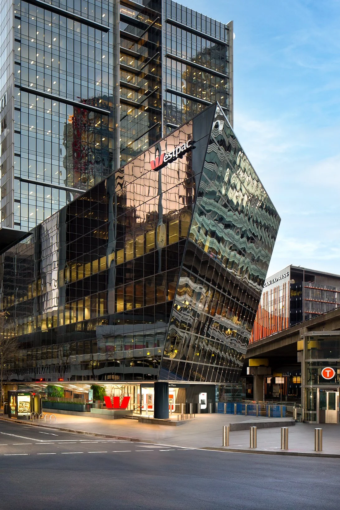 Modern glass office building with the Westpac logo, located in an urban downtown area during dusk. The building features an angular, reflective façade with multiple floors visible through large windows.
