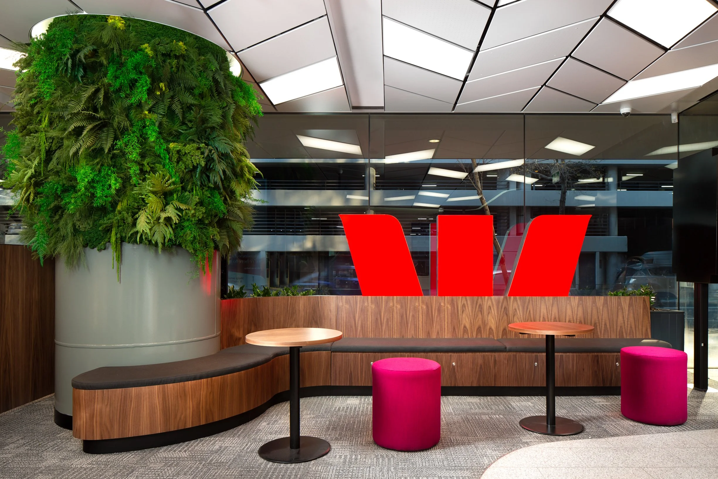 Interior of a modern office or lobby with a green wall, wooden seating area, round tables, and pink ottomans, with a backlit ceiling and glass walls.