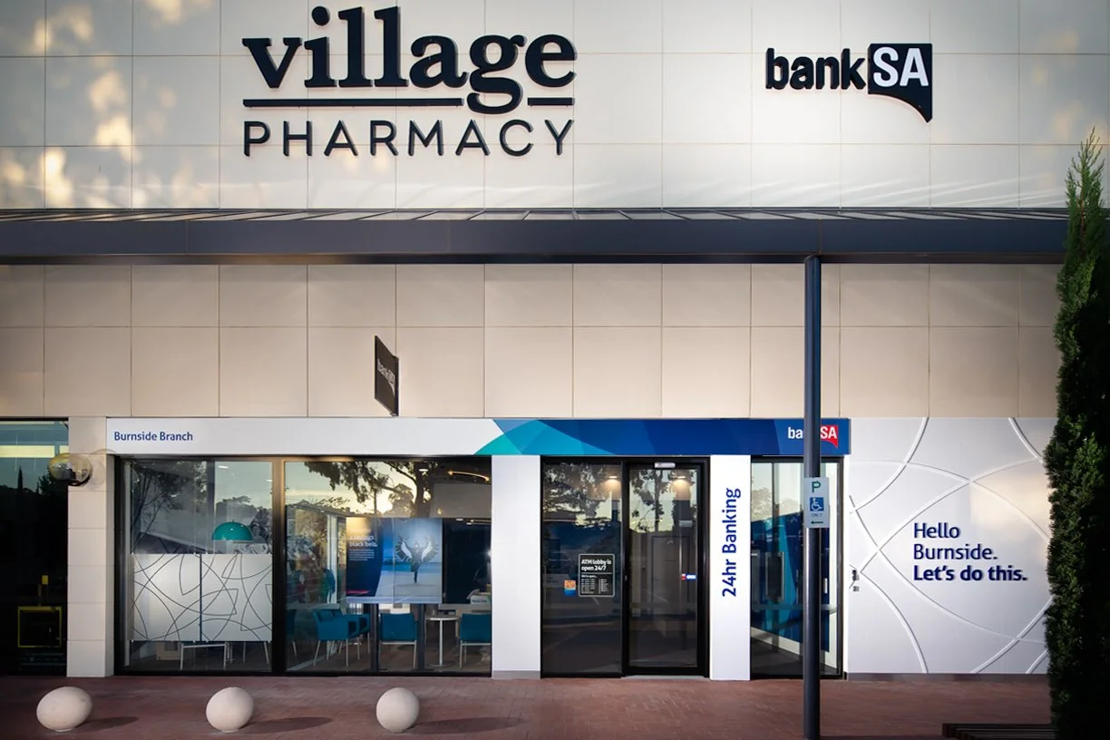 Exterior view of a pharmacy and bank branch with signage reading 'Village Pharmacy' and 'Bank SA'. The pharmacy has glass doors and windows with a seating area visible inside. There is a sign indicating 24-hour banking services next to the entrance, 