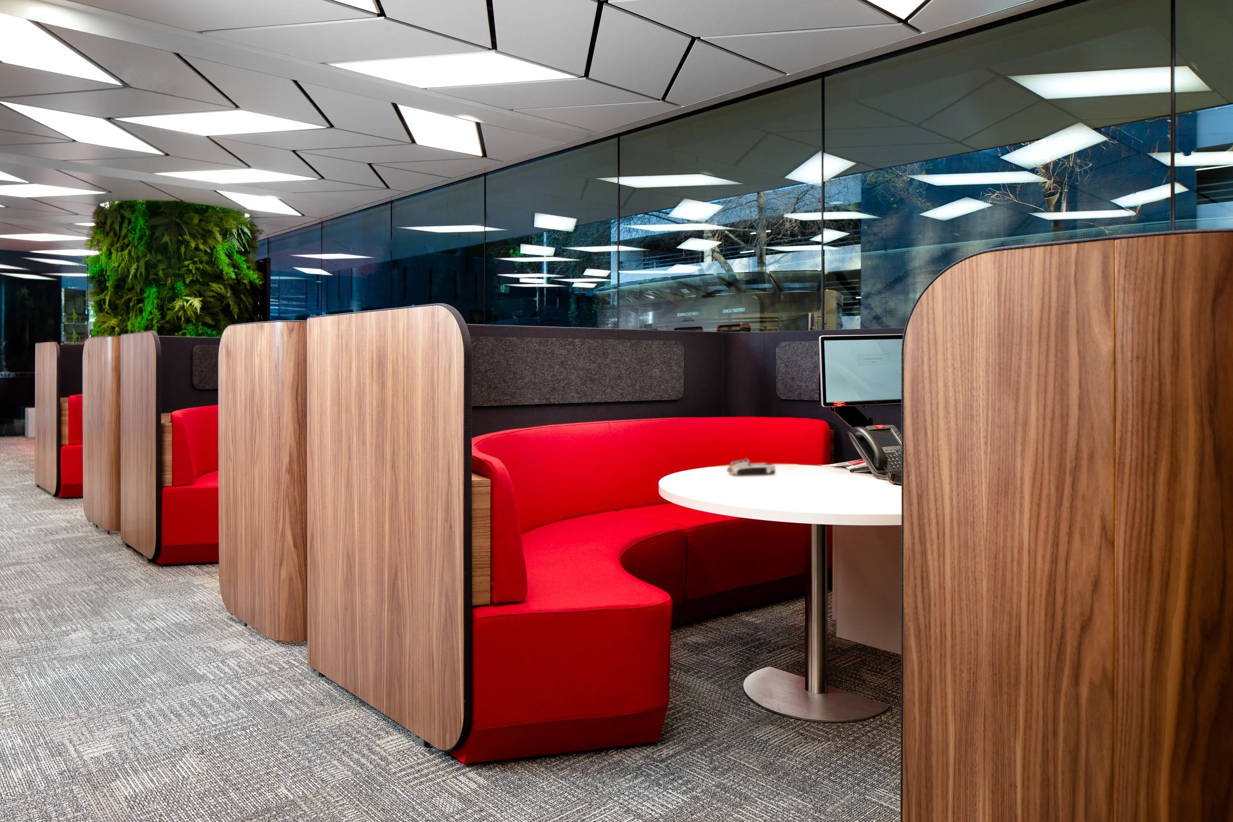 Modern office lounge with semi-private booths, red cushioned seating, wooden dividers, a desk with a telephone, and a large window reflecting ceiling lights and outdoor trees.