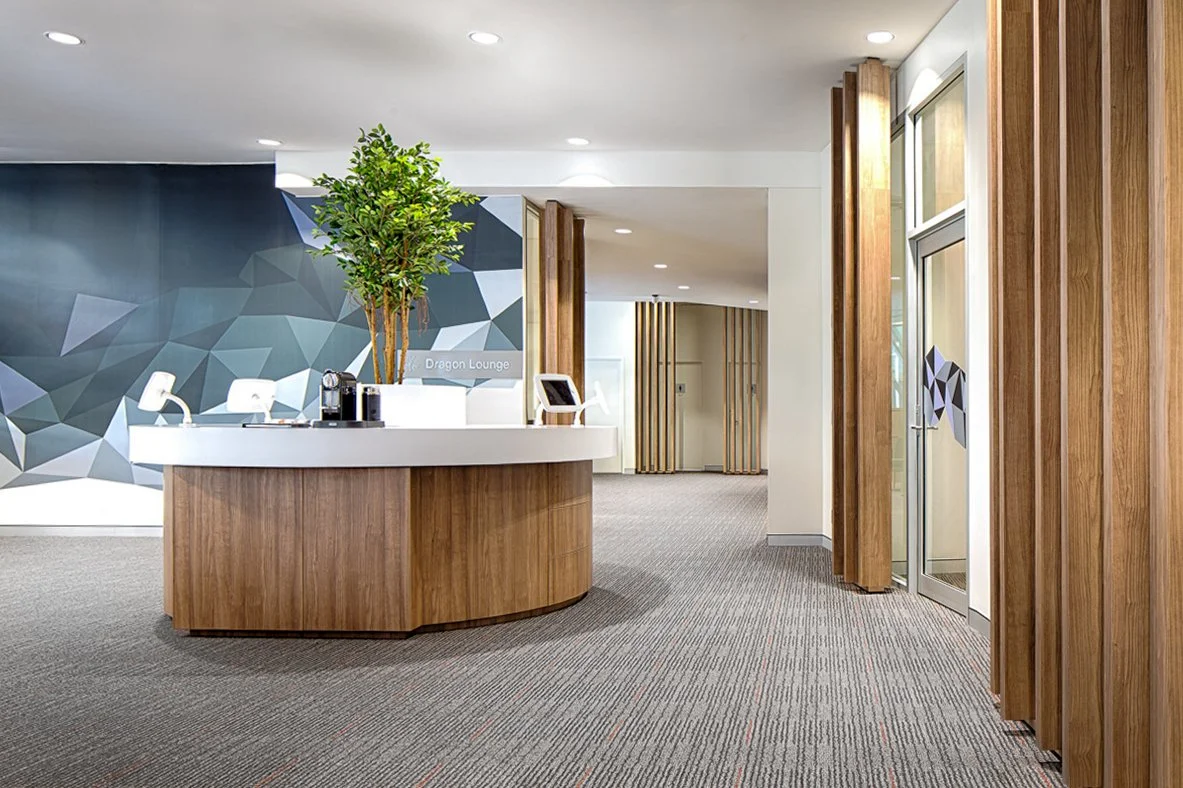 Hotel reception area with a round wooden desk, a potted plant, and a geometric wall design in the background.