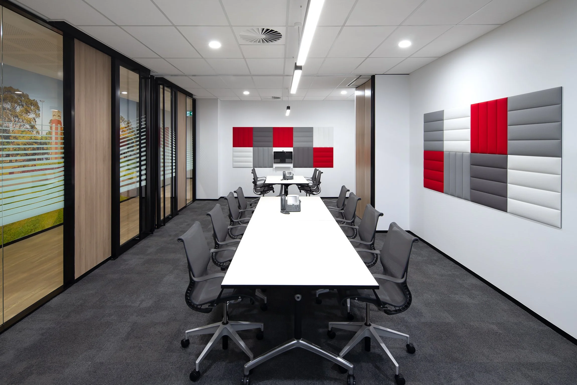 A modern conference room with a long white table surrounded by gray chairs, a wall with red, white, and gray acoustic panels, glass walls with blinds, and a ceiling with recessed lighting and a linear light fixture.