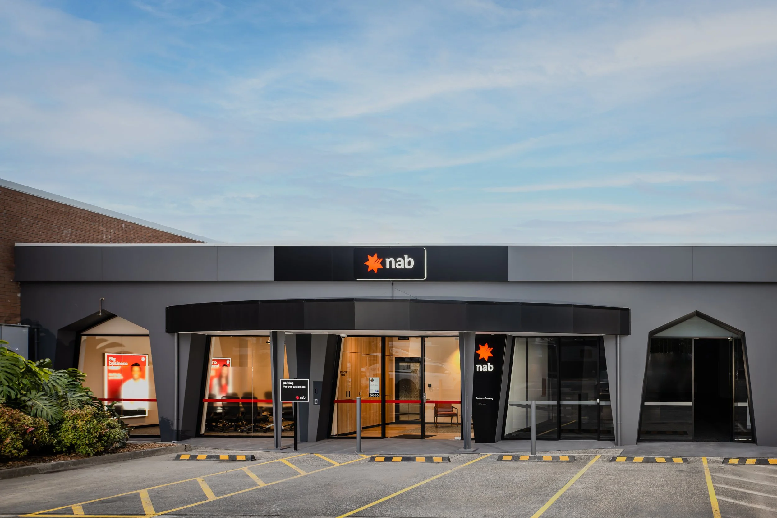 Front view of a NAB bank branch with a modern design, large glass windows, and a sign with the NAB logo above the entrance. There are parking spaces in front, some signage, and greenery on the side.