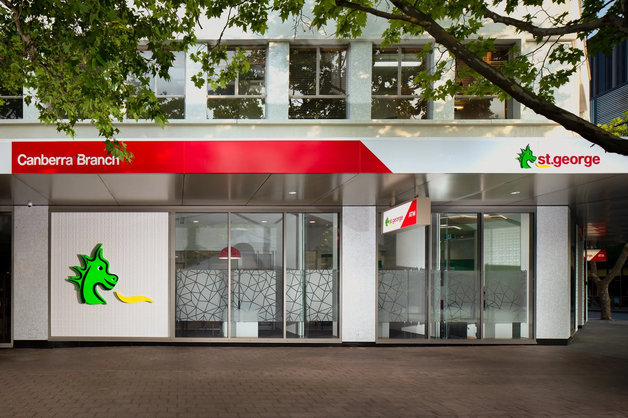 Storefront of Canberra Branch and St. George bank with glass windows, red and white signage, and a green dragon logo, partially obscured by tree branches.