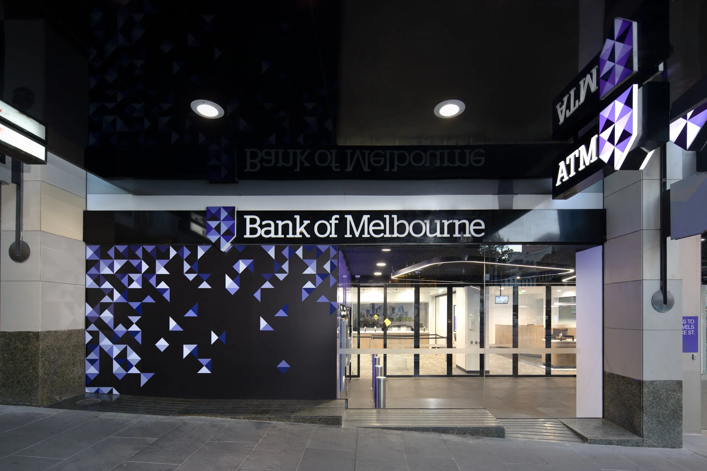 Facade of a modern bank branch with illuminated signs reading 'Bank of Melbourne' and 'ATM', featuring geometric purple and black designs on the exterior wall, and an entrance with automatic sliding glass doors.