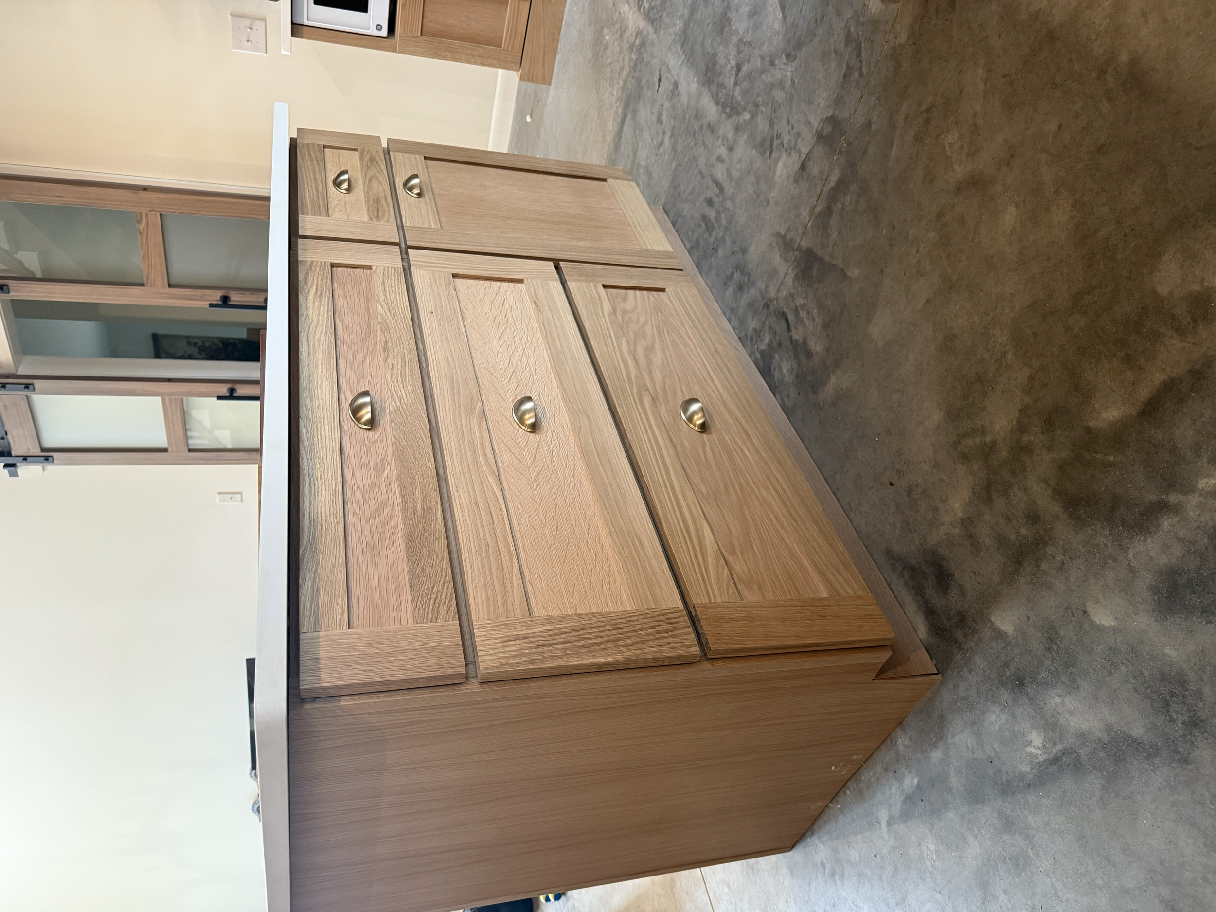 A wooden kitchen cabinet with multiple drawers and brass handles, situated on a concrete floor in a room with beige walls.