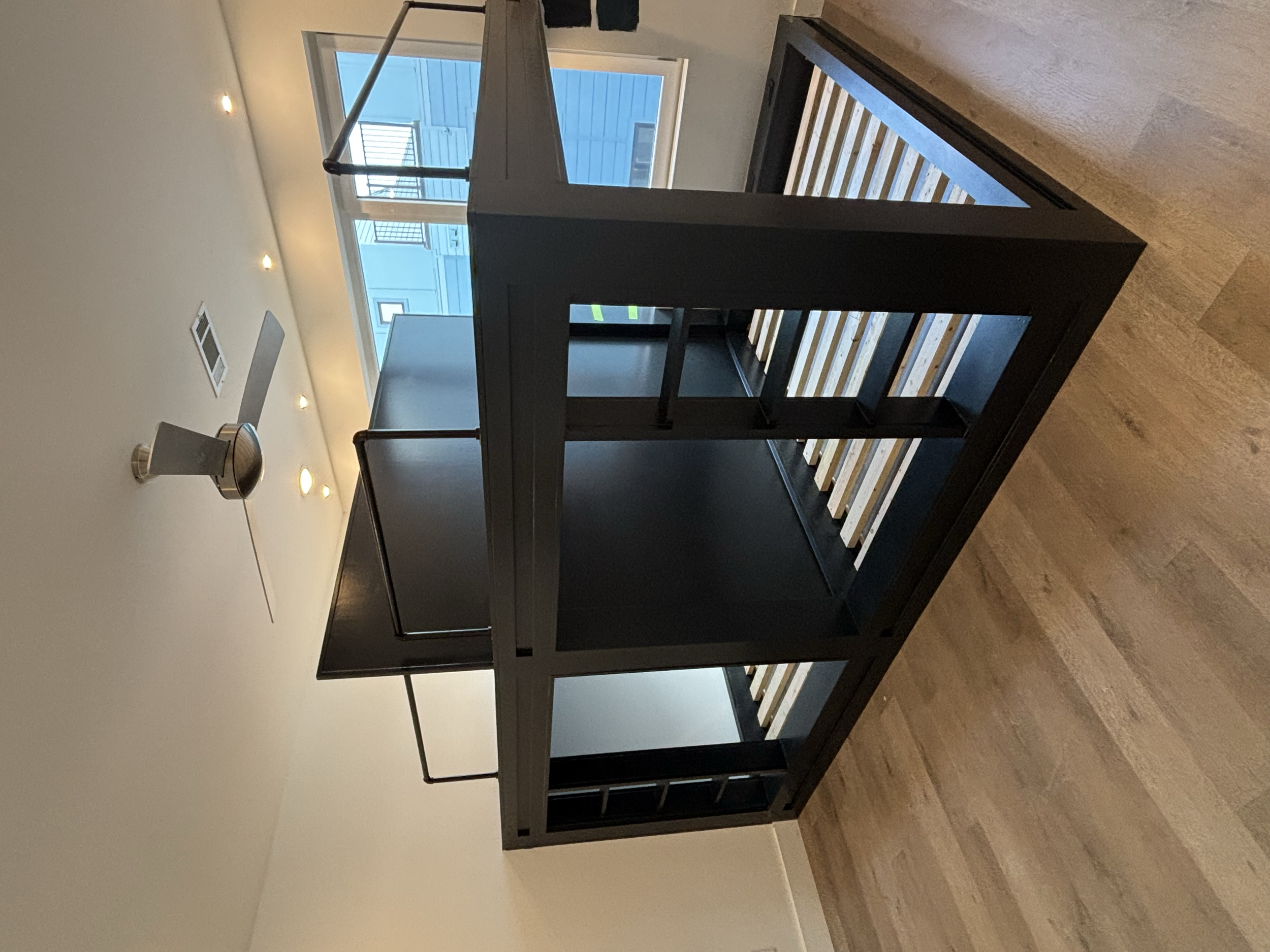Interior of a room showing a dark wooden bed frame, a large window with a balcony outside, and ceiling lights.