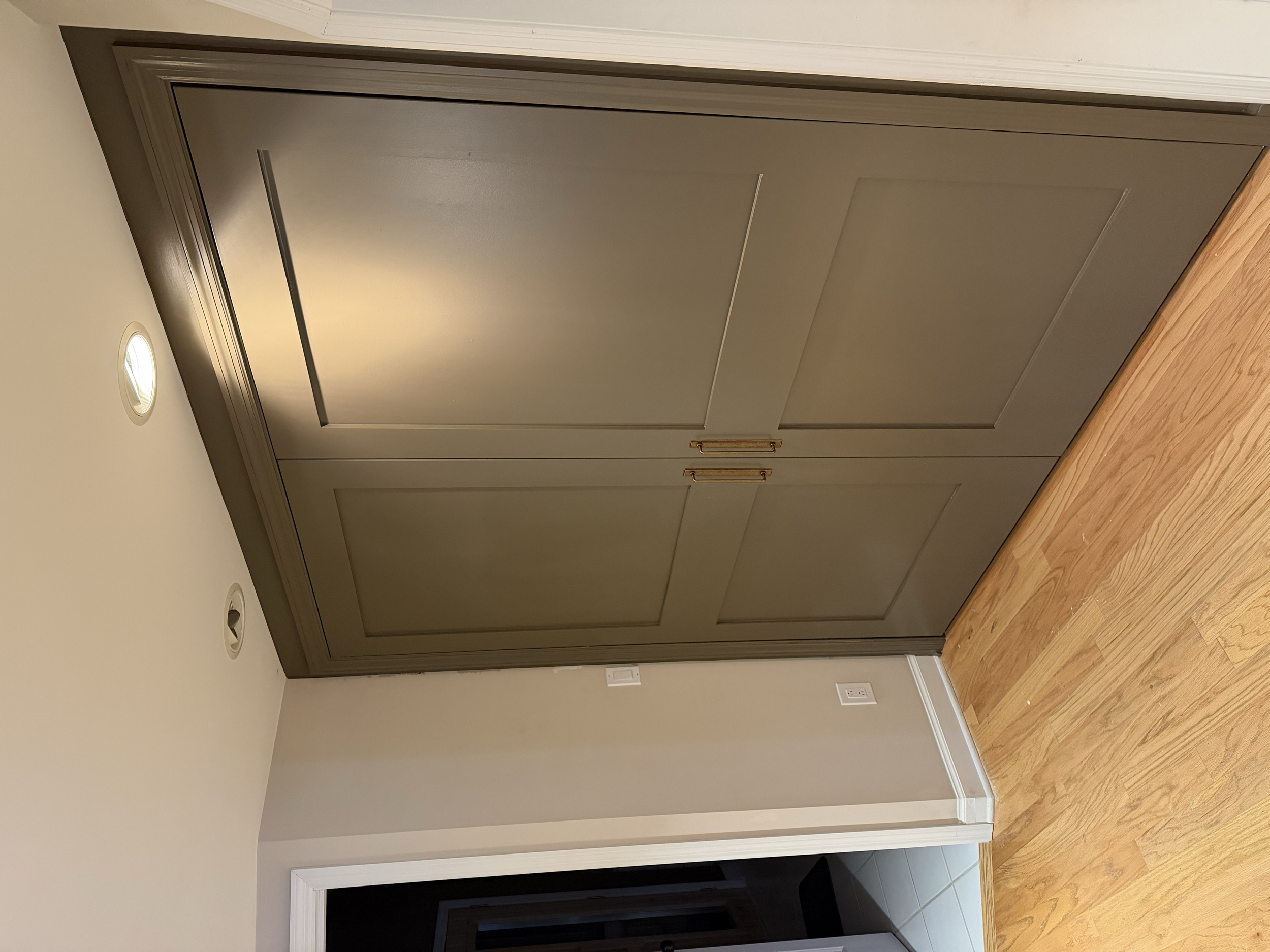 A closed gray door with a metallic handle, set in a white door frame. The door is in a room with hardwood flooring, white wall, and ceiling with built-in lighting.