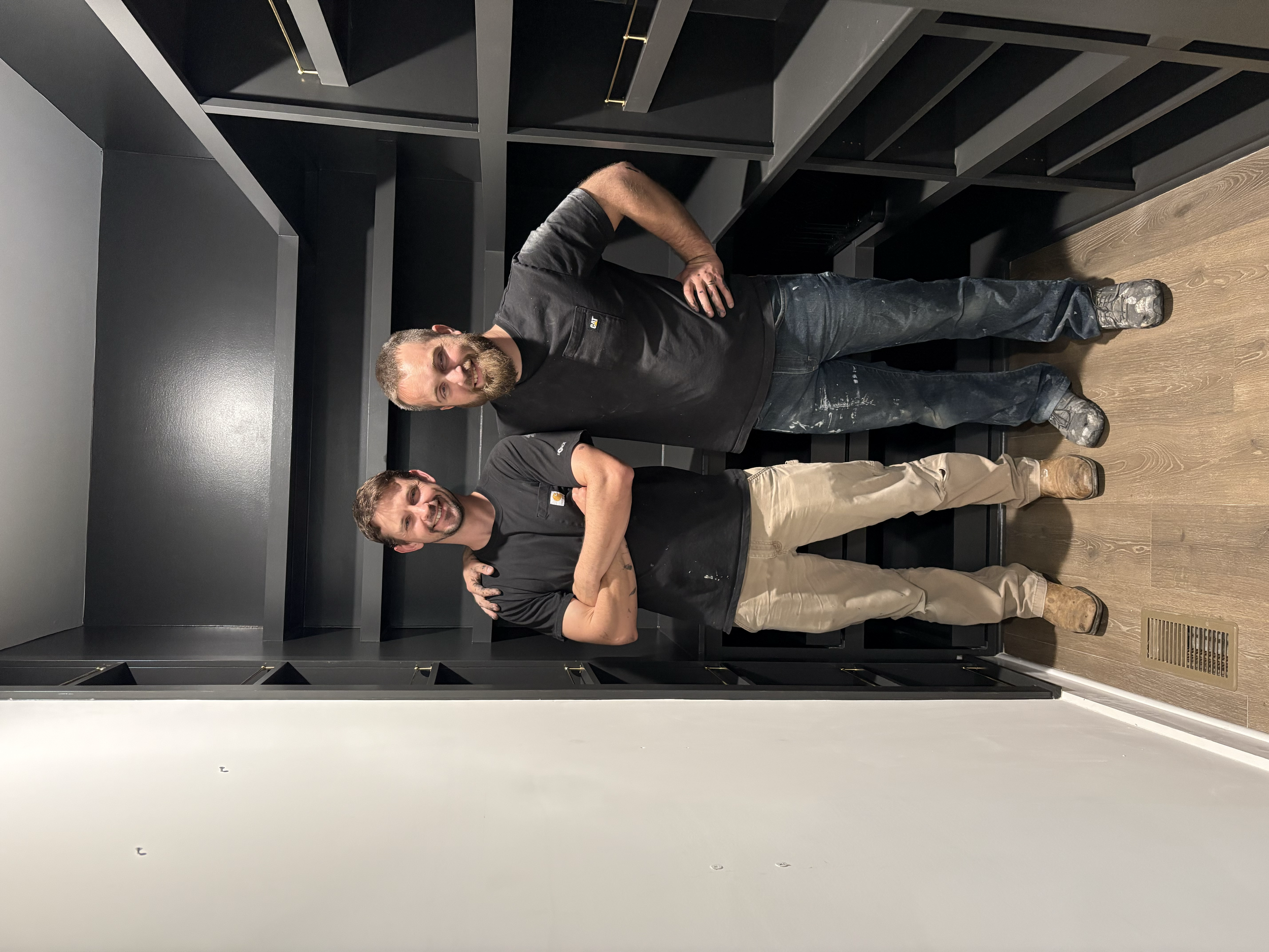 Two men standing side by side in an elevator with black interior. They are smiling, one with a beard and the other clean-shaven. Both are wearing black t-shirts and jeans. One man has his arm around the other's shoulder.