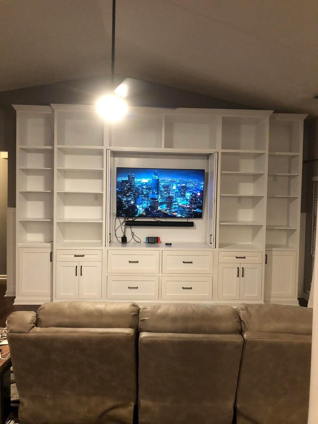 Living room with white built-in entertainment center, a flat-screen TV showing a cityscape at night, and a beige sofa in the foreground.