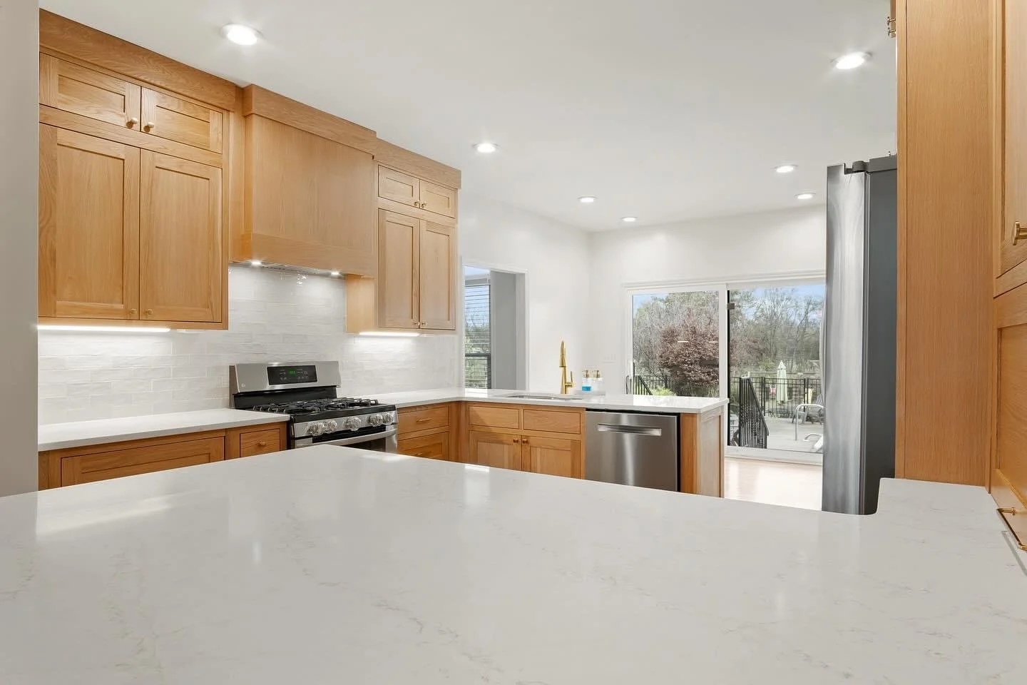 Modern kitchen with wooden cabinets, white countertops, stainless steel appliances, and a sliding glass door leading to an outdoor patio.
