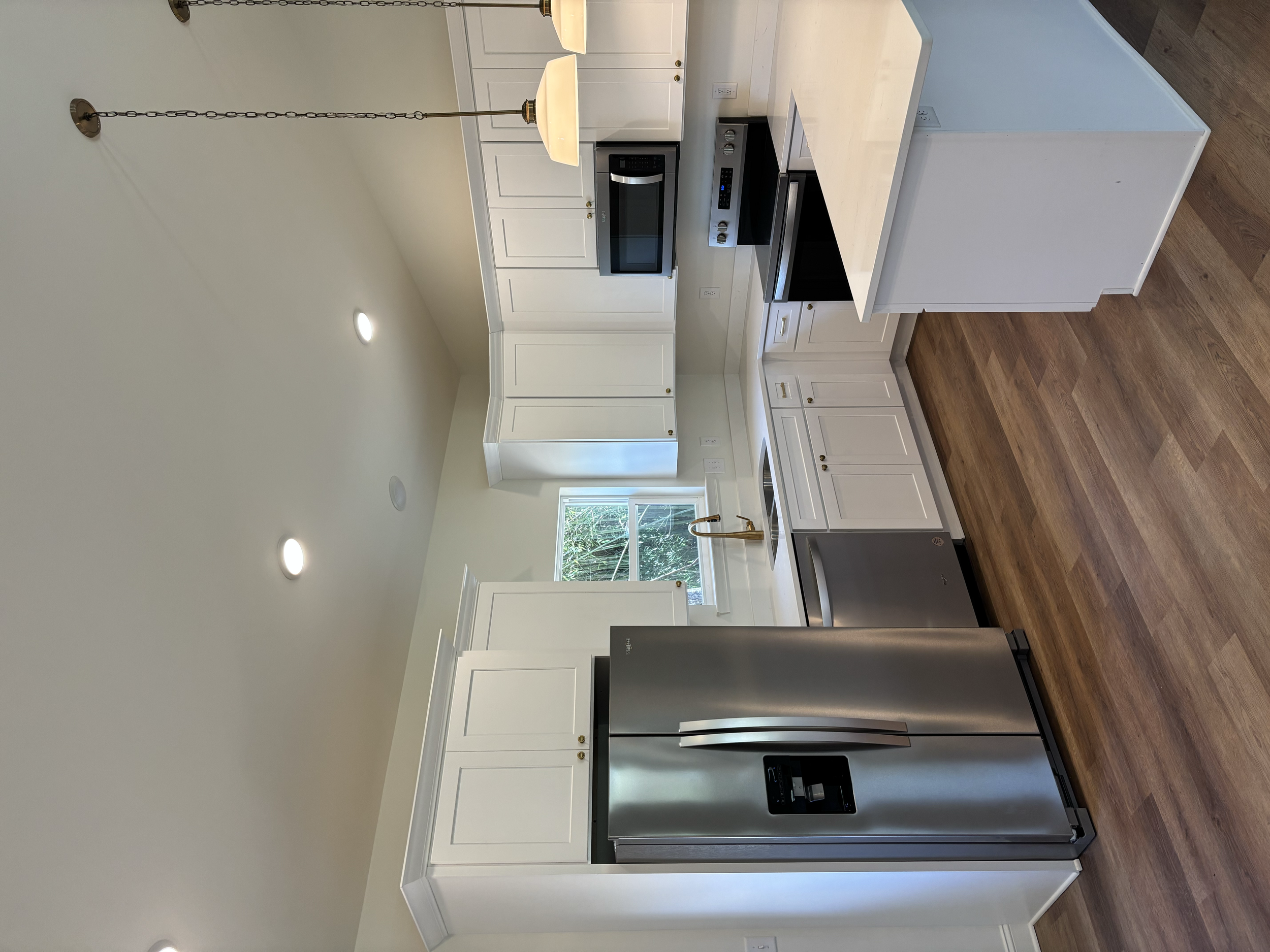 A modern kitchen with white cabinetry, a stainless steel refrigerator, a microwave, a stove, and a white marble countertop with a gold-colored faucet. There are recessed lights in the ceiling and a window showing greenery outside.