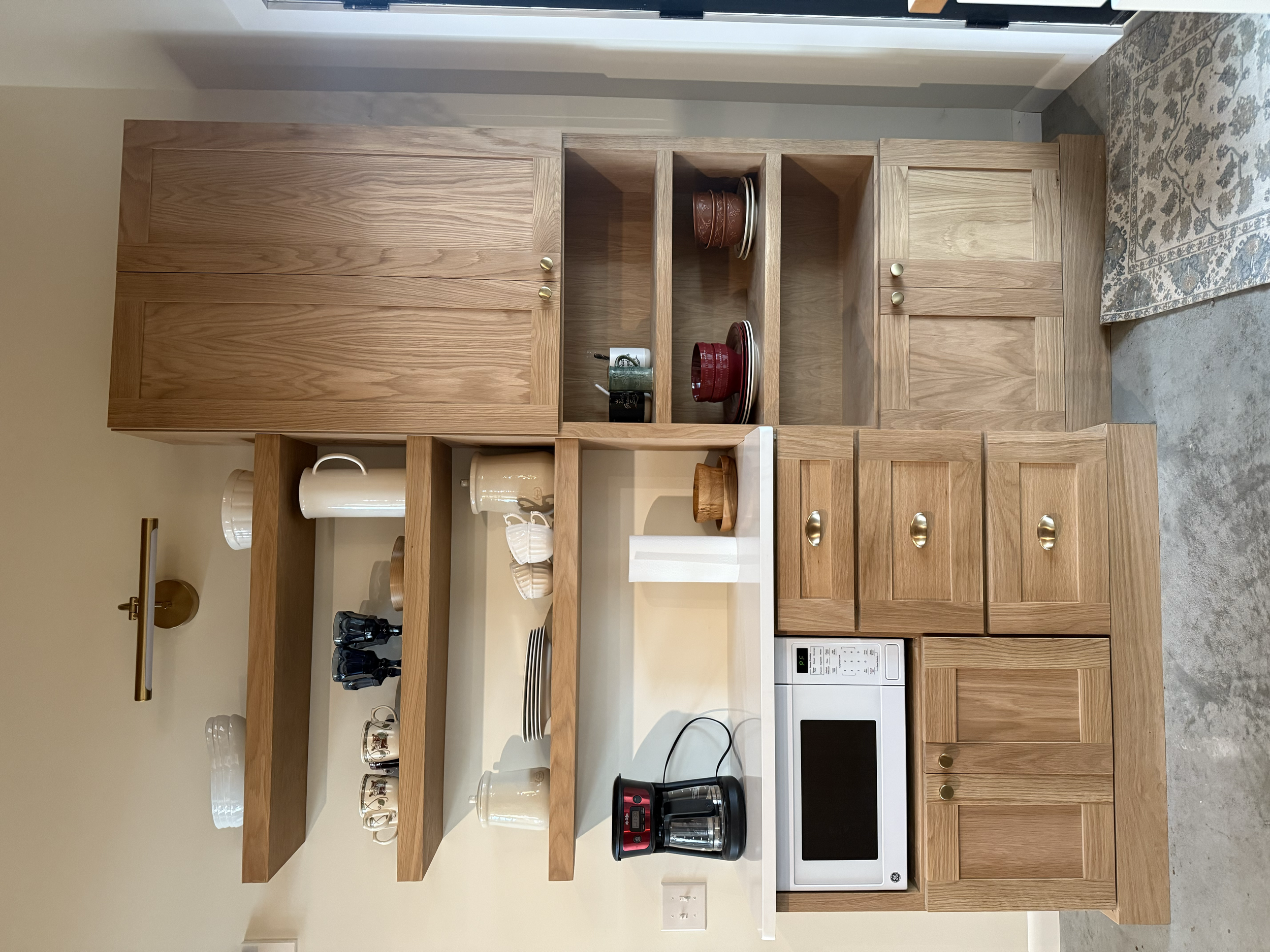 A wooden kitchen cabinet and shelves filled with dishes, mugs, and kitchen items. The bottom has a microwave and a coffee maker. The kitchen has a concrete floor and a beige wall.