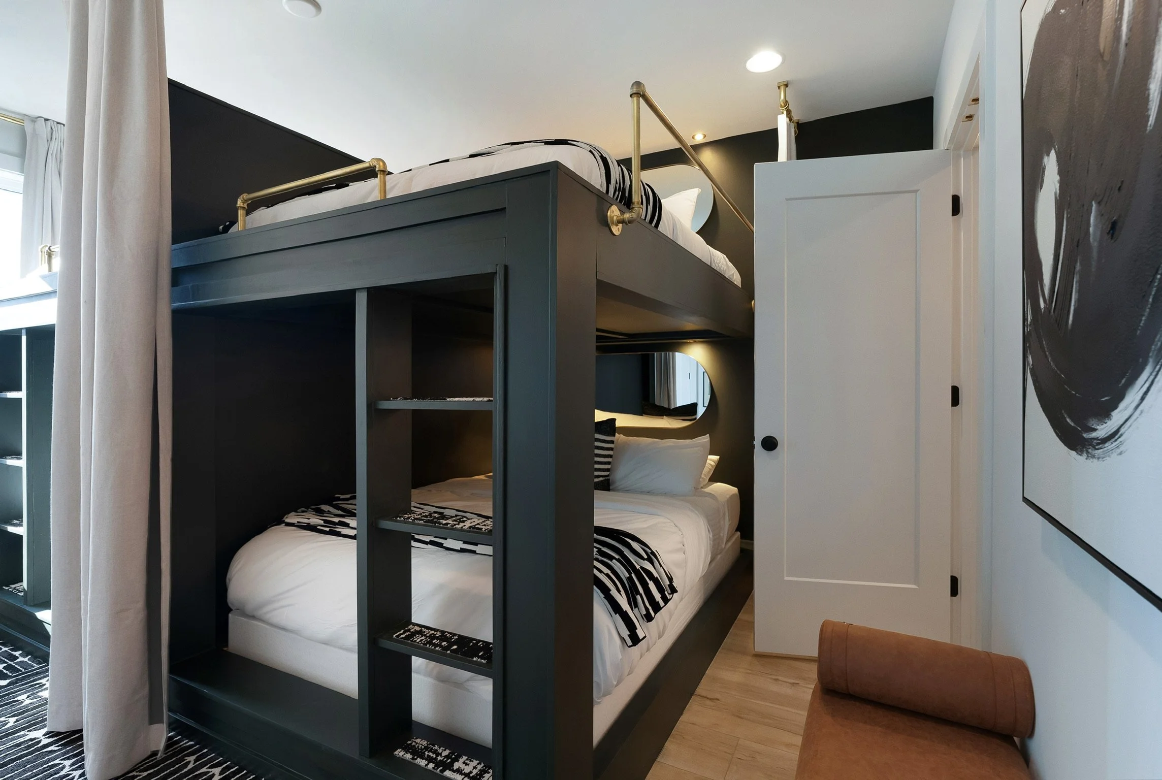 A modern bedroom with a bunk bed featuring a black and white bedding set. The upper bunk has brass railings, and the lower bunk has pillows and bedding. There is a white door and abstract artwork on the walls. A small brown chair is in the foreground