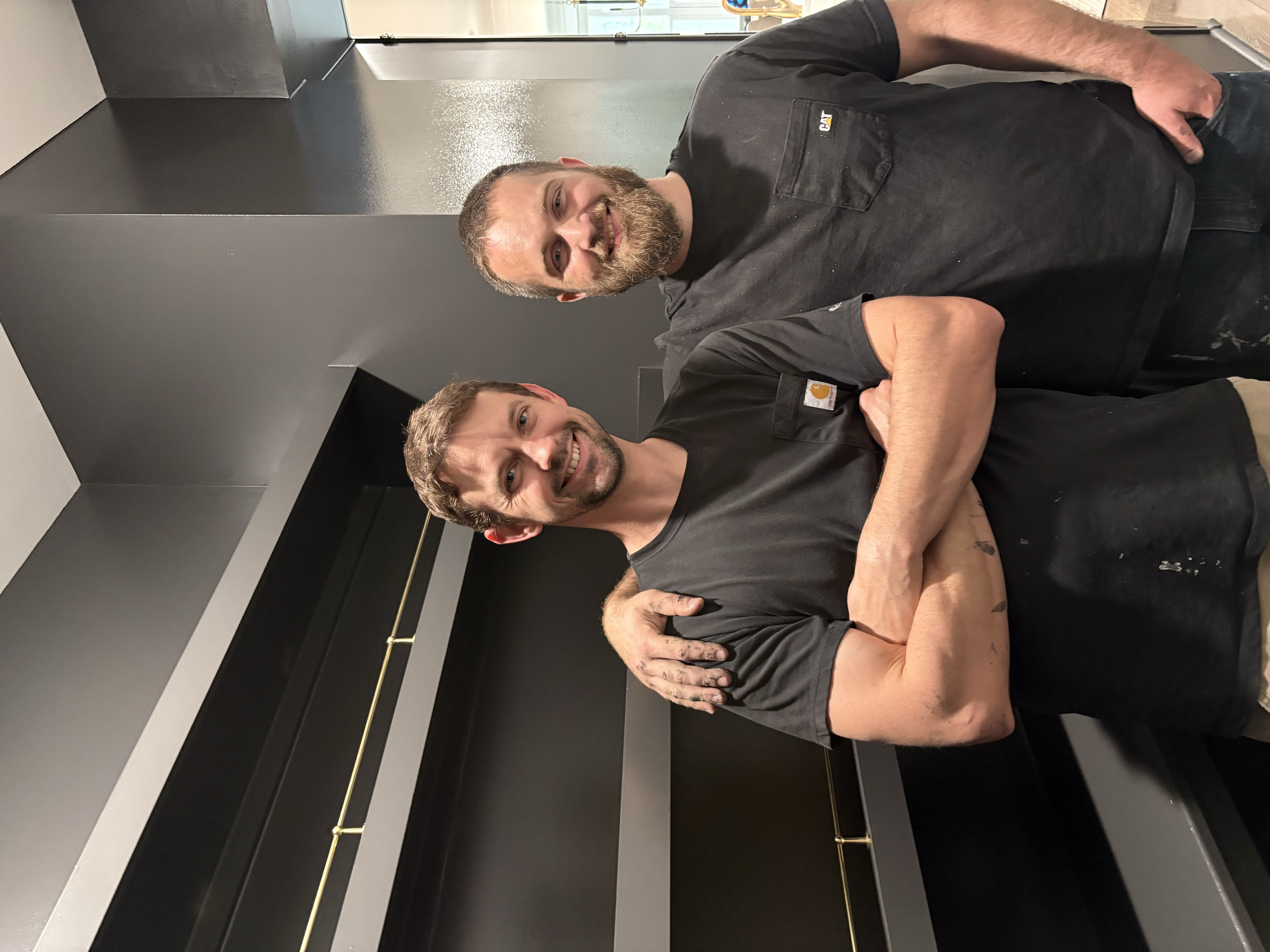 Two smiling men wearing black shirts, standing close together with arms around each other's shoulders in a modern indoor setting.