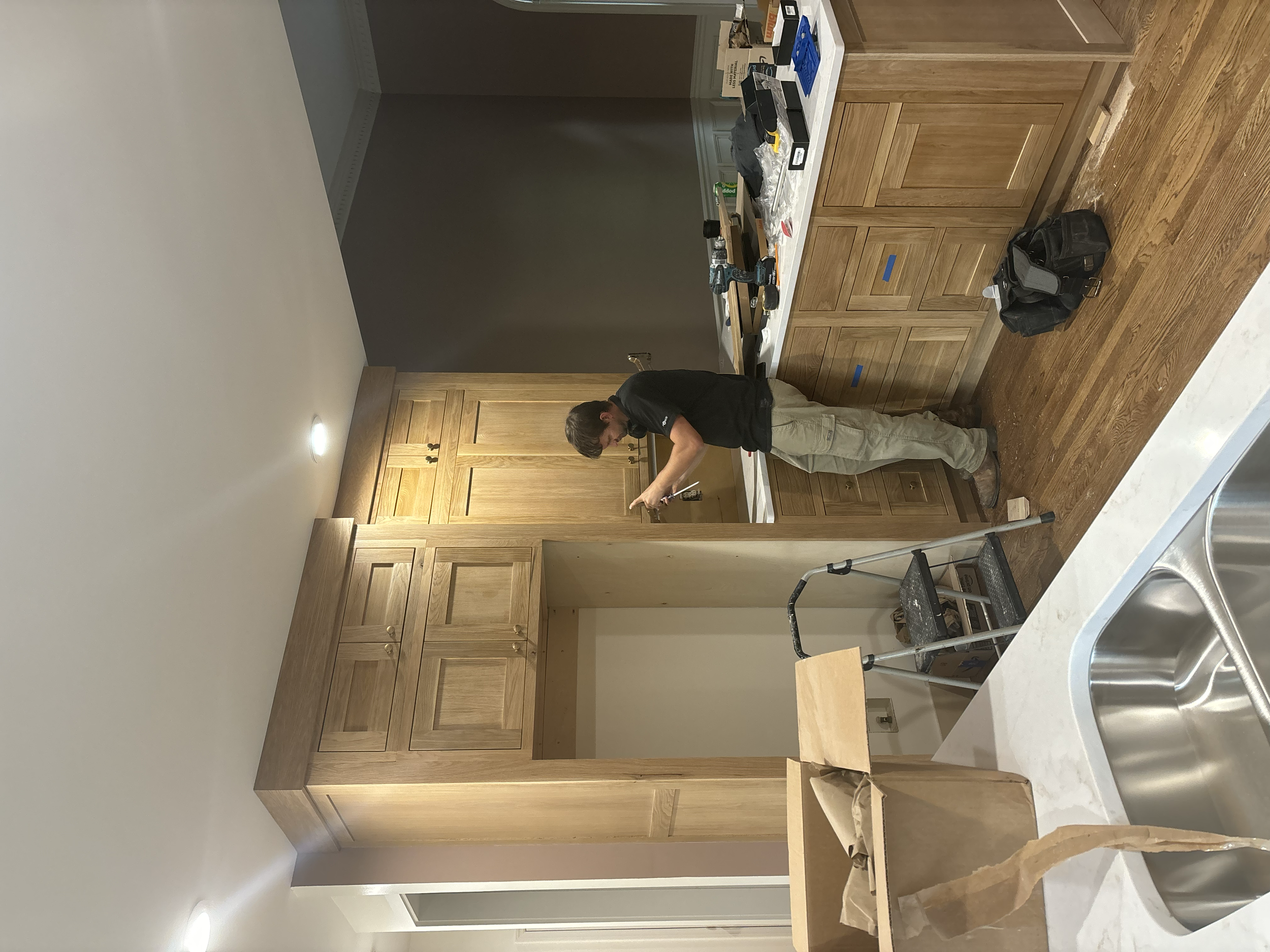 A person working on installing or building kitchen cabinets in a kitchen with wooden cabinetry and a white countertop. There's a step ladder, tools, and materials on the counter.