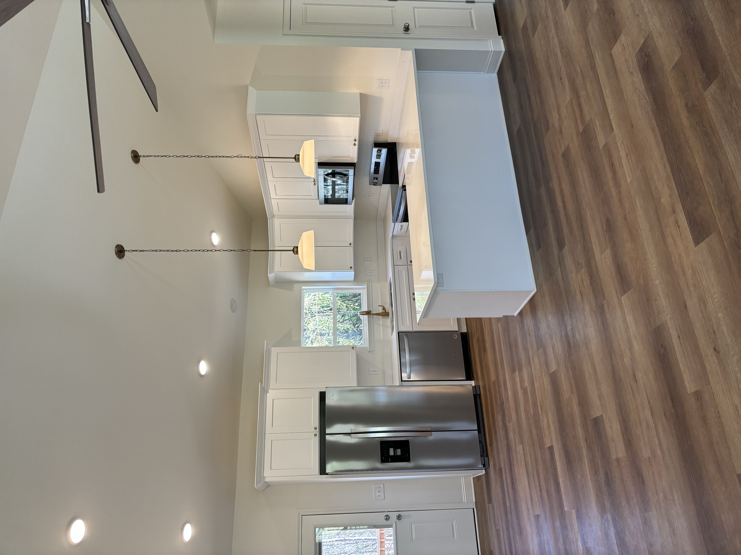 View of a modern kitchen with white cabinets, a stainless steel refrigerator, built-in oven, microwave, and wooden flooring, illuminated by ceiling lights and natural light through windows.