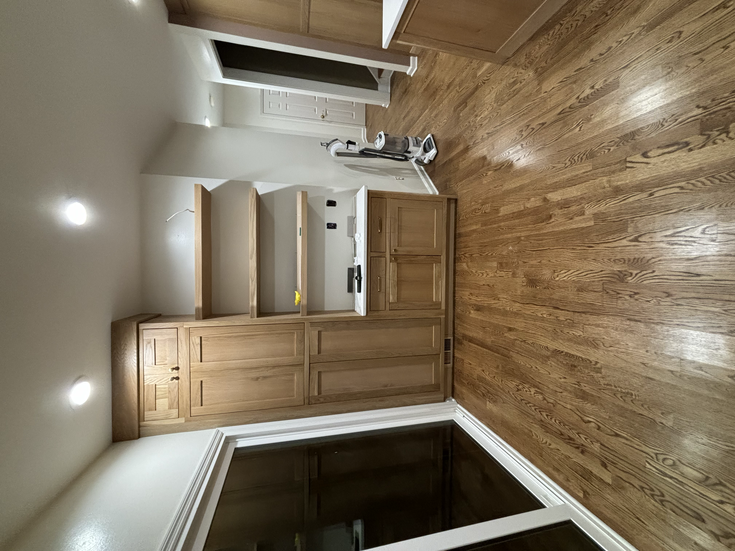 Interior of a room with wooden flooring, a wooden cabinet, and shelves. There are recessed ceiling lights and a vacuum cleaner in the corner.