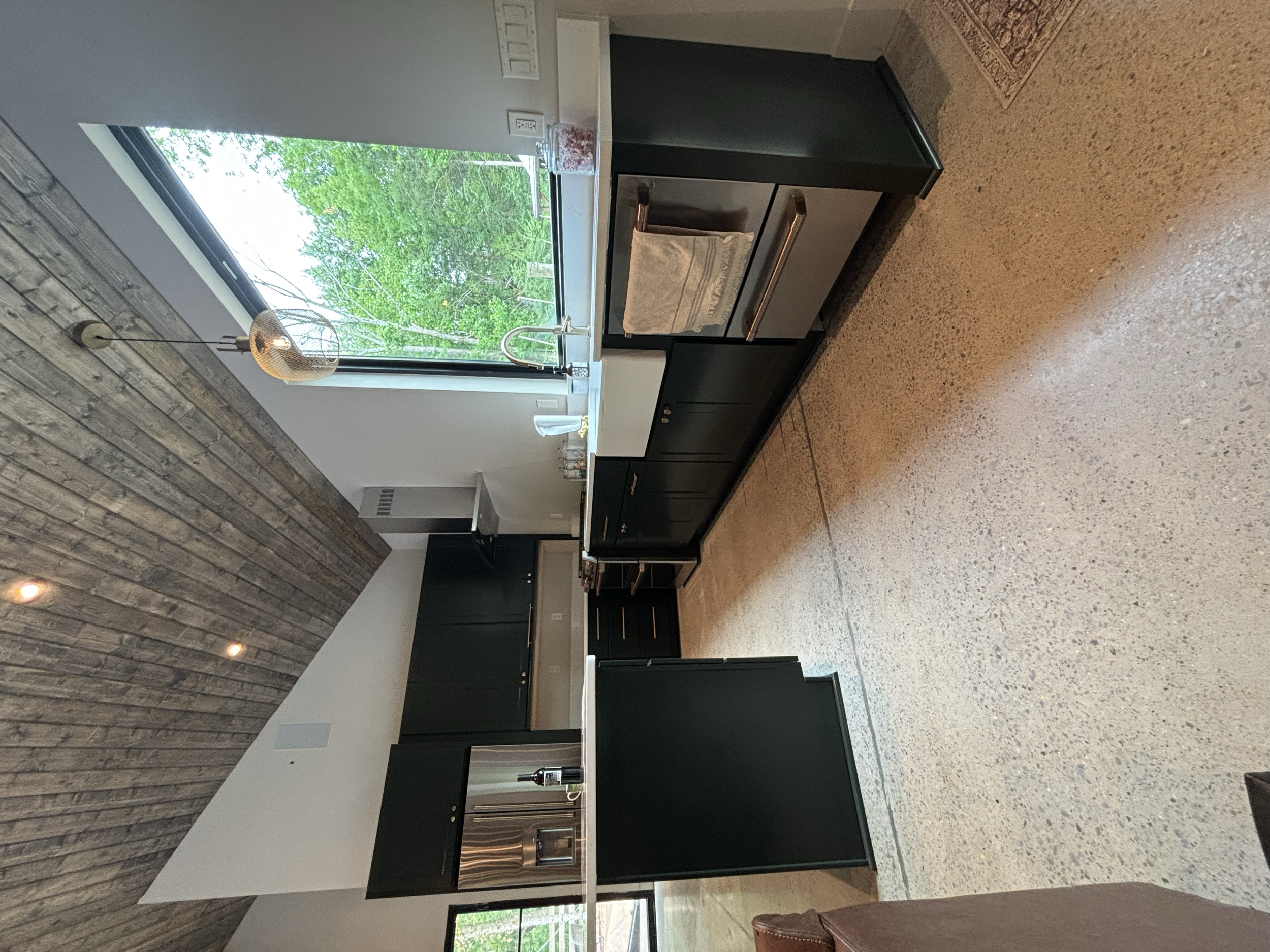 A modern kitchen with black cabinetry, a large window showing green trees outside, a stone wall, and a pendant light hanging from the ceiling.
