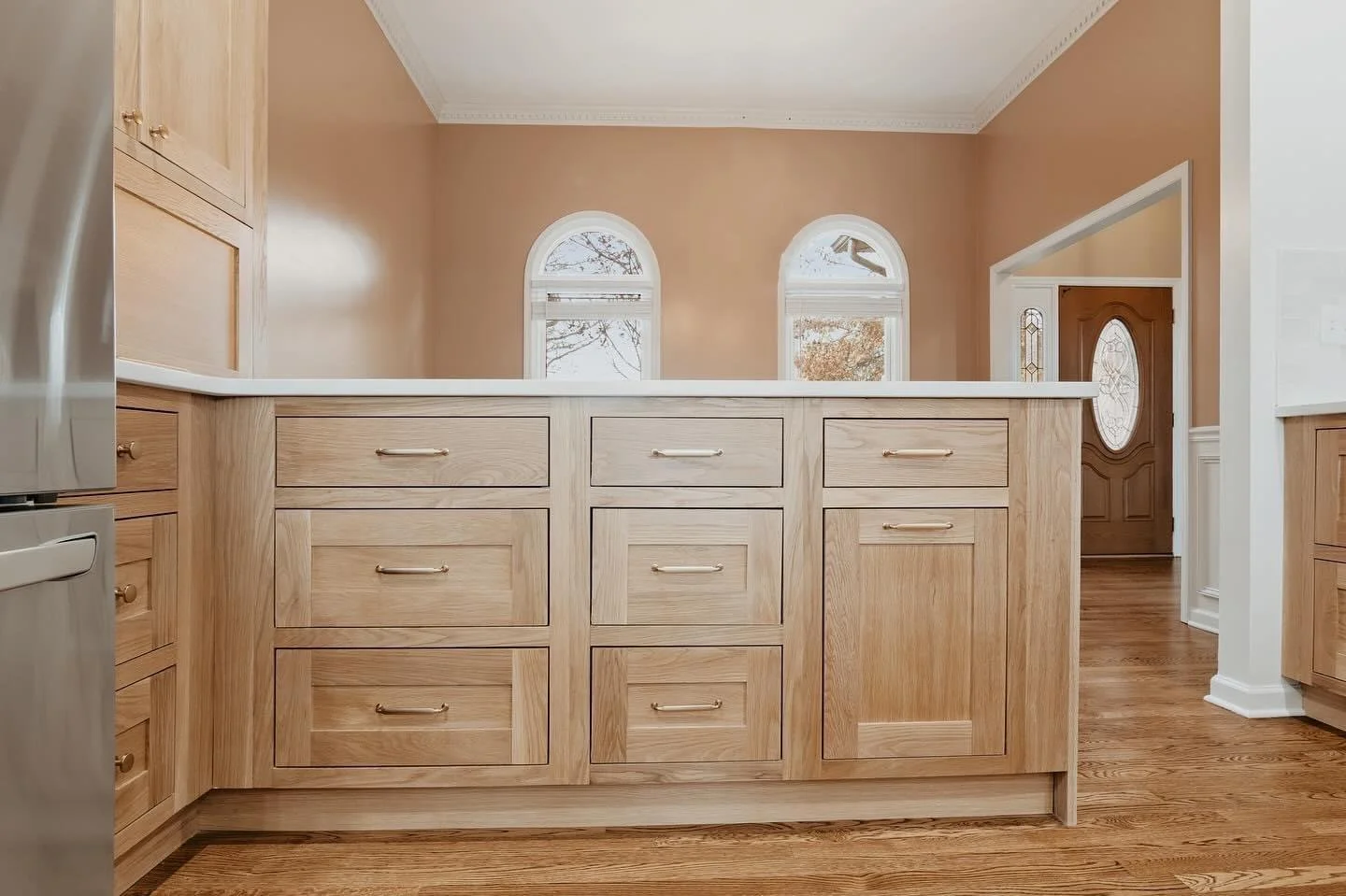 A kitchen island with wooden drawers and a countertop, hardwood floors, and beige walls with two arched windows and a front door in the background.