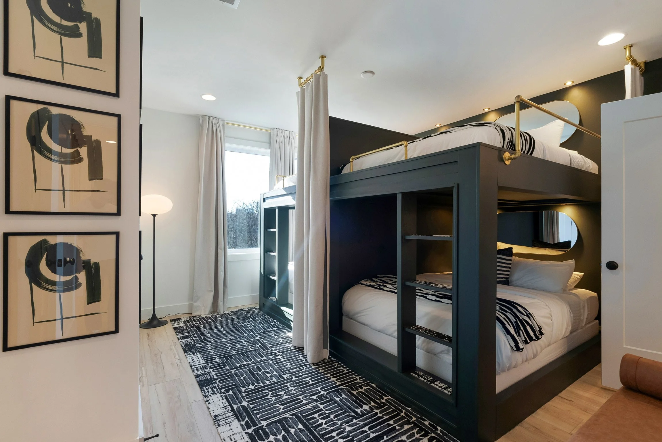 A room with bunk beds, a window with white curtains, and wall art featuring abstract black and beige designs. There's a lamp to the left and a rug with black and white patterns on the floor.