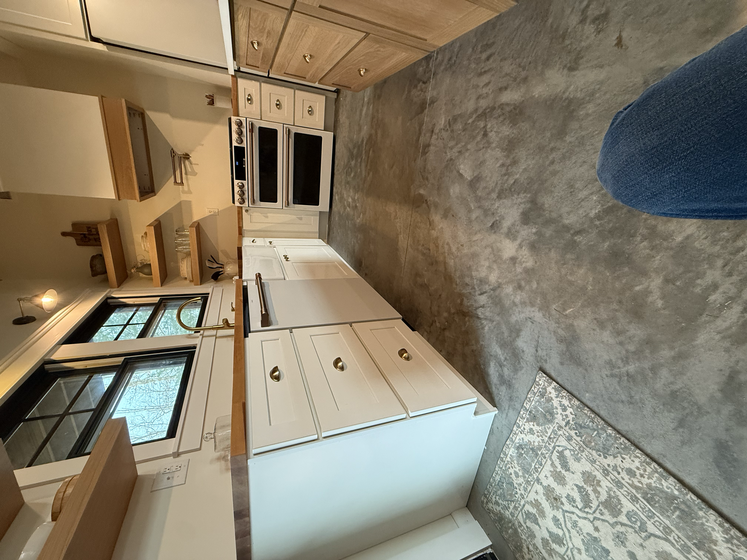 A kitchen with white cabinets, a wooden countertop, a black stove, and large windows. The floor is made of gray concrete, and part of a blue patterned rug is visible. There is a blue denim pant on the right side.