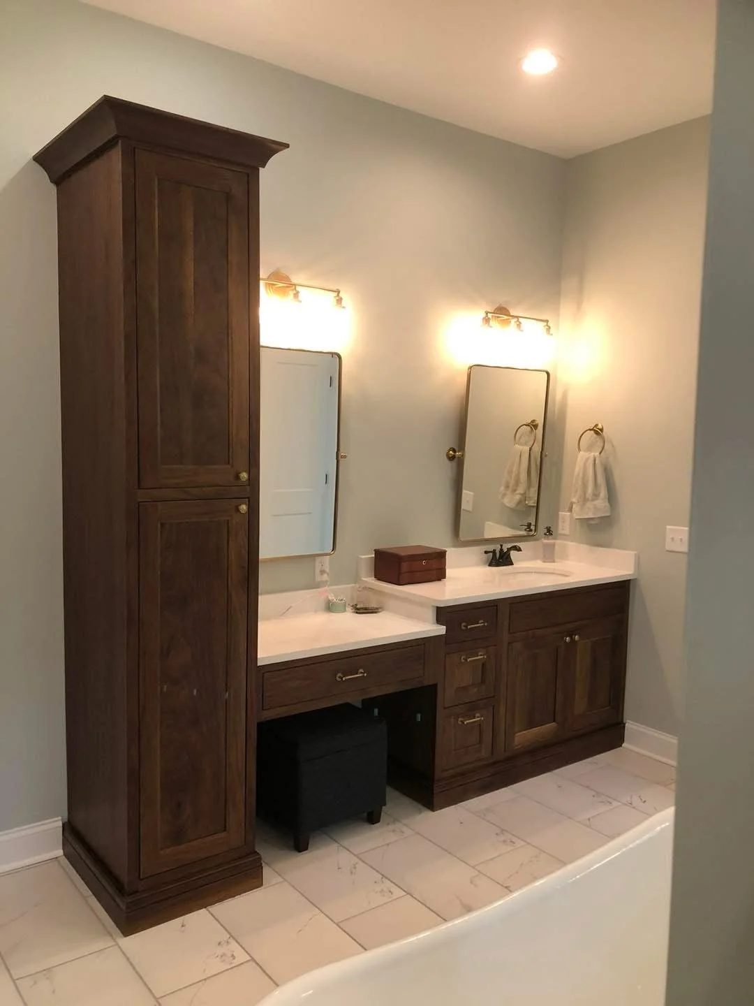 Bathroom vanity with two mirrors, wooden cabinetry, and a tall storage cabinet to the left. There are two wall-mounted lights above the mirrors, and a small black stool under the left side of the vanity. The floor is tiled and there are two towels ha