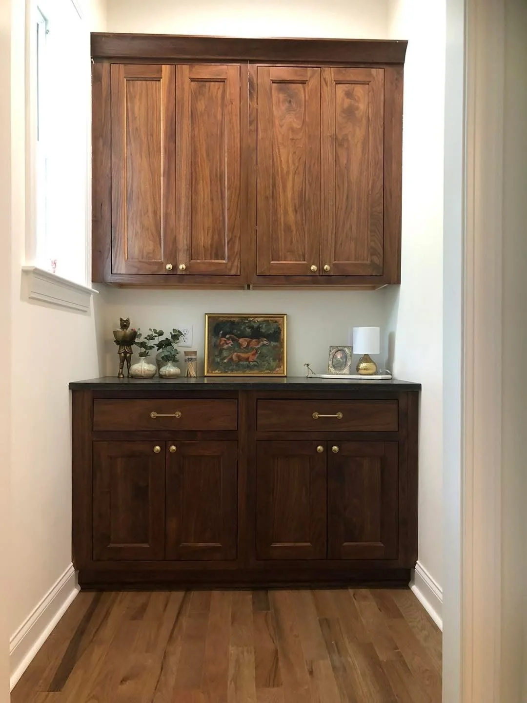 A wooden cabinet with upper and lower sections, decorated with small plants, a painting, a table lamp, and picture frames, in a well-lit room.