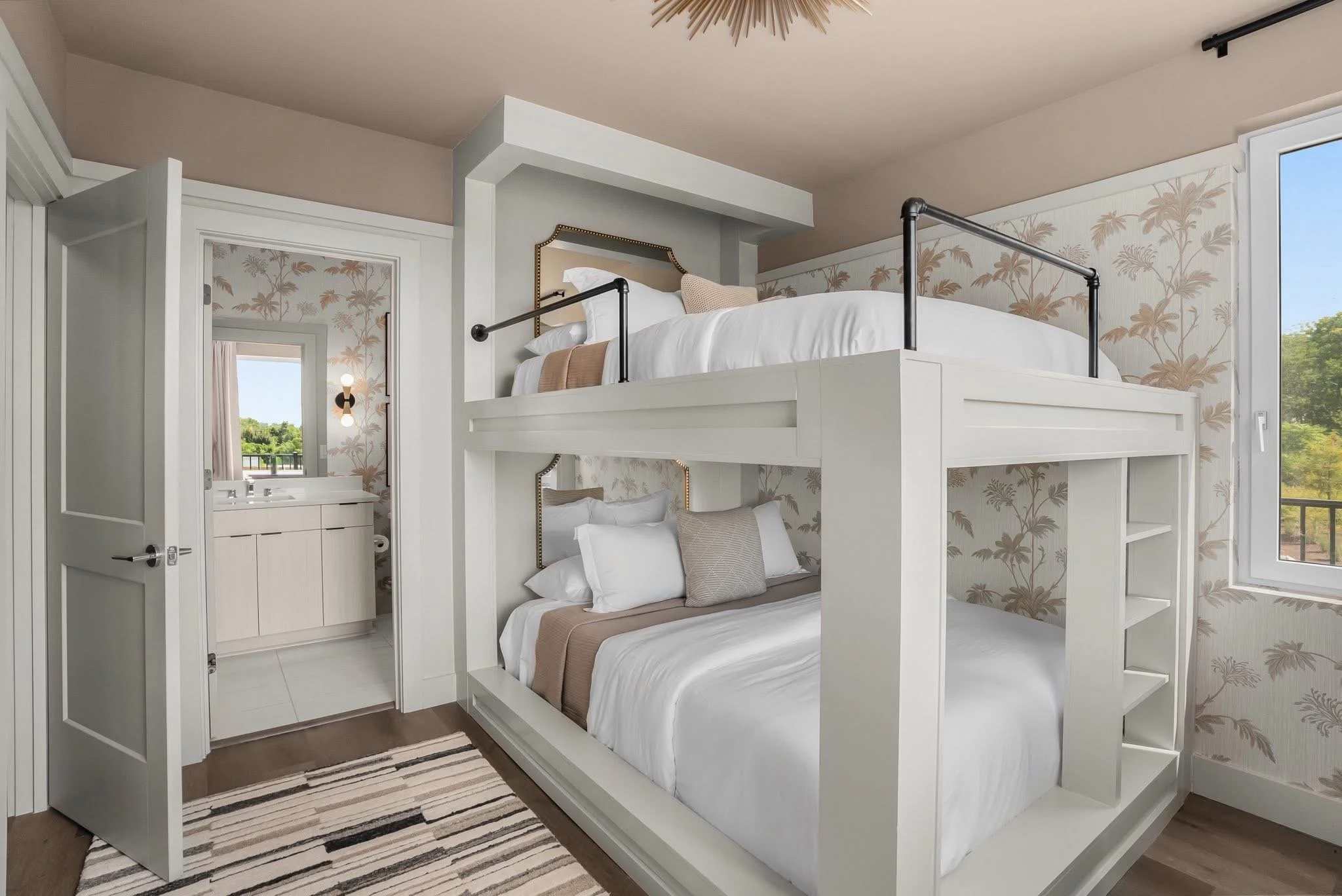 A bedroom featuring a white bunk bed with black metal guardrails, a beige pillow, and white bedding. The room has floral wallpaper, a window showing greenery outside, a small mirror with a vanity, and a striped rug on the wooden floor.