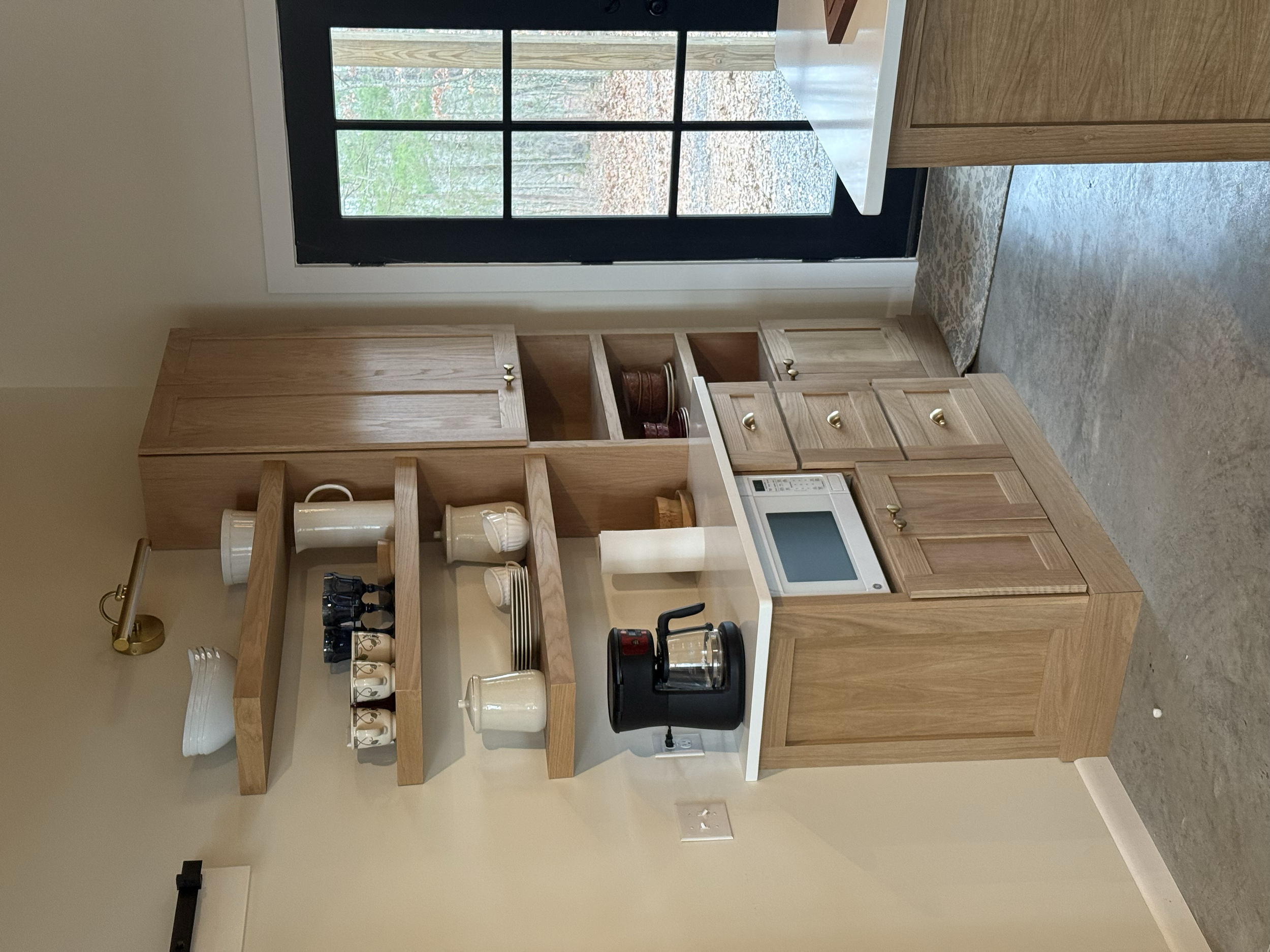 A kitchen with wooden cabinets, open shelves with dishes, a microwave, a coffee maker, and a window showing a view of trees outside.