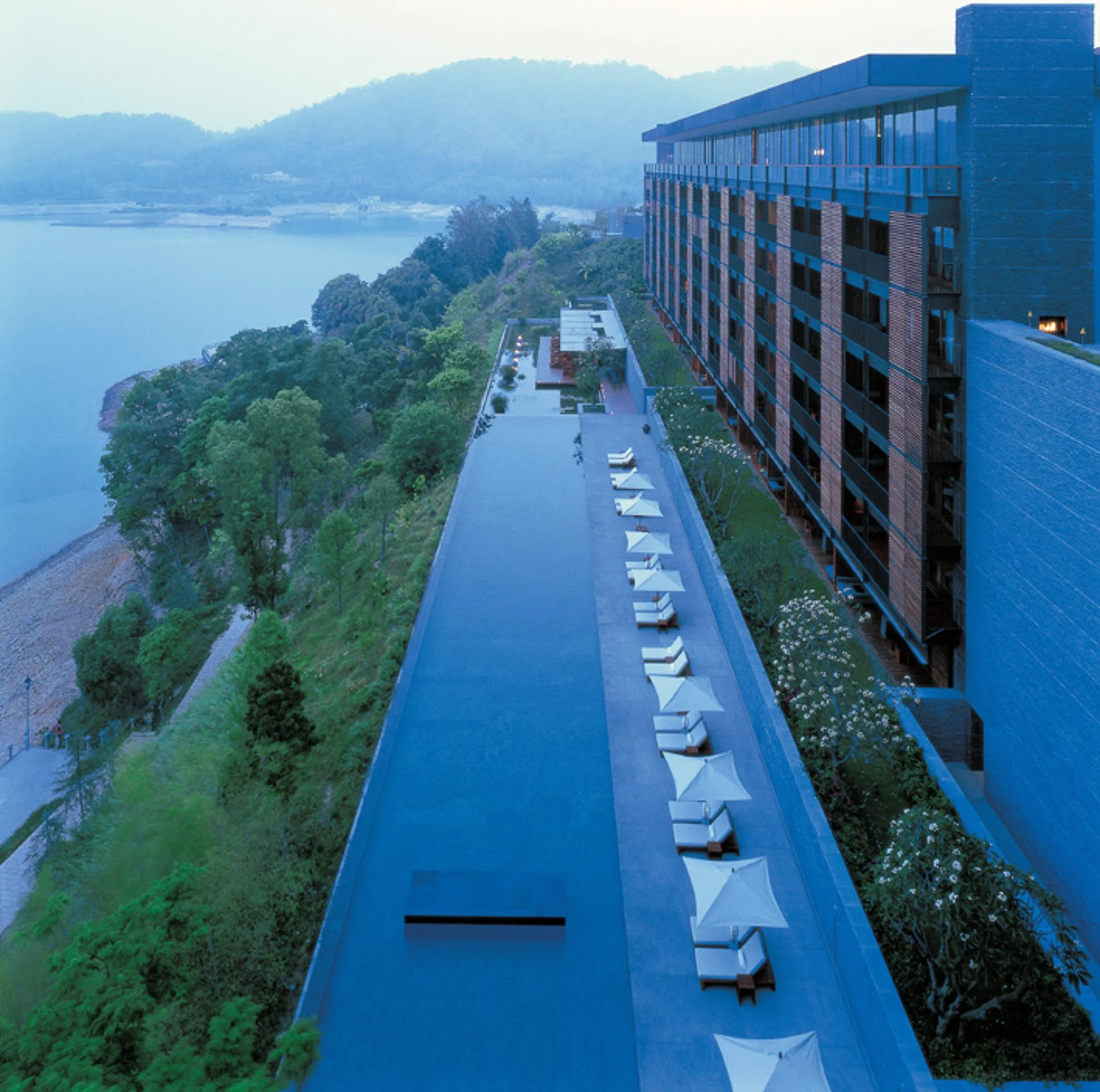 Modern multi-story building with large windows, balconies, and a rooftop terrace situated on a hillside overlooking a body of water with trees and mountains in the background, during daylight.